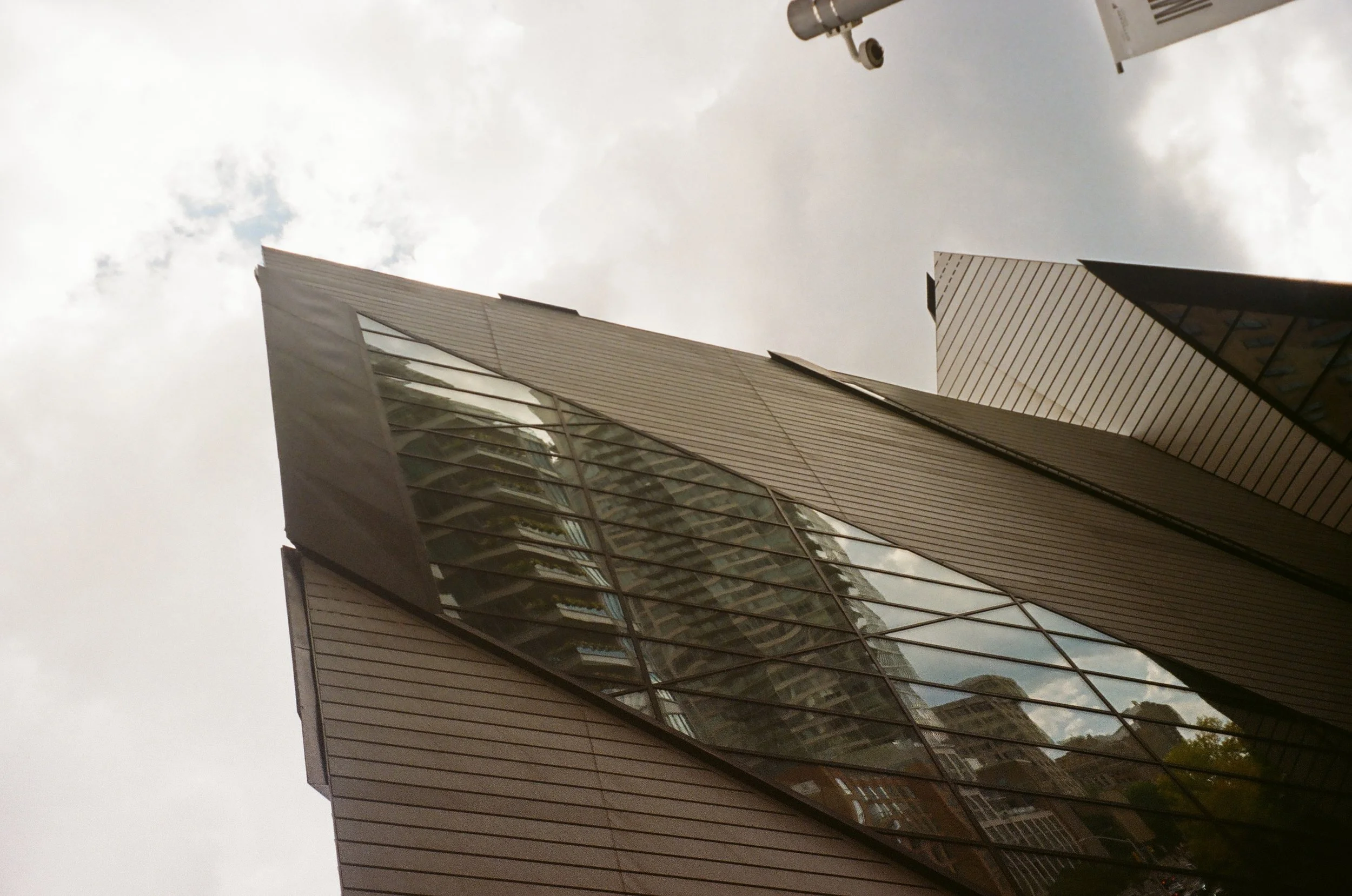 Modern high-rise building with angled glass and metal facade, reflecting nearby structures and partly cloudy sky, viewed from below.