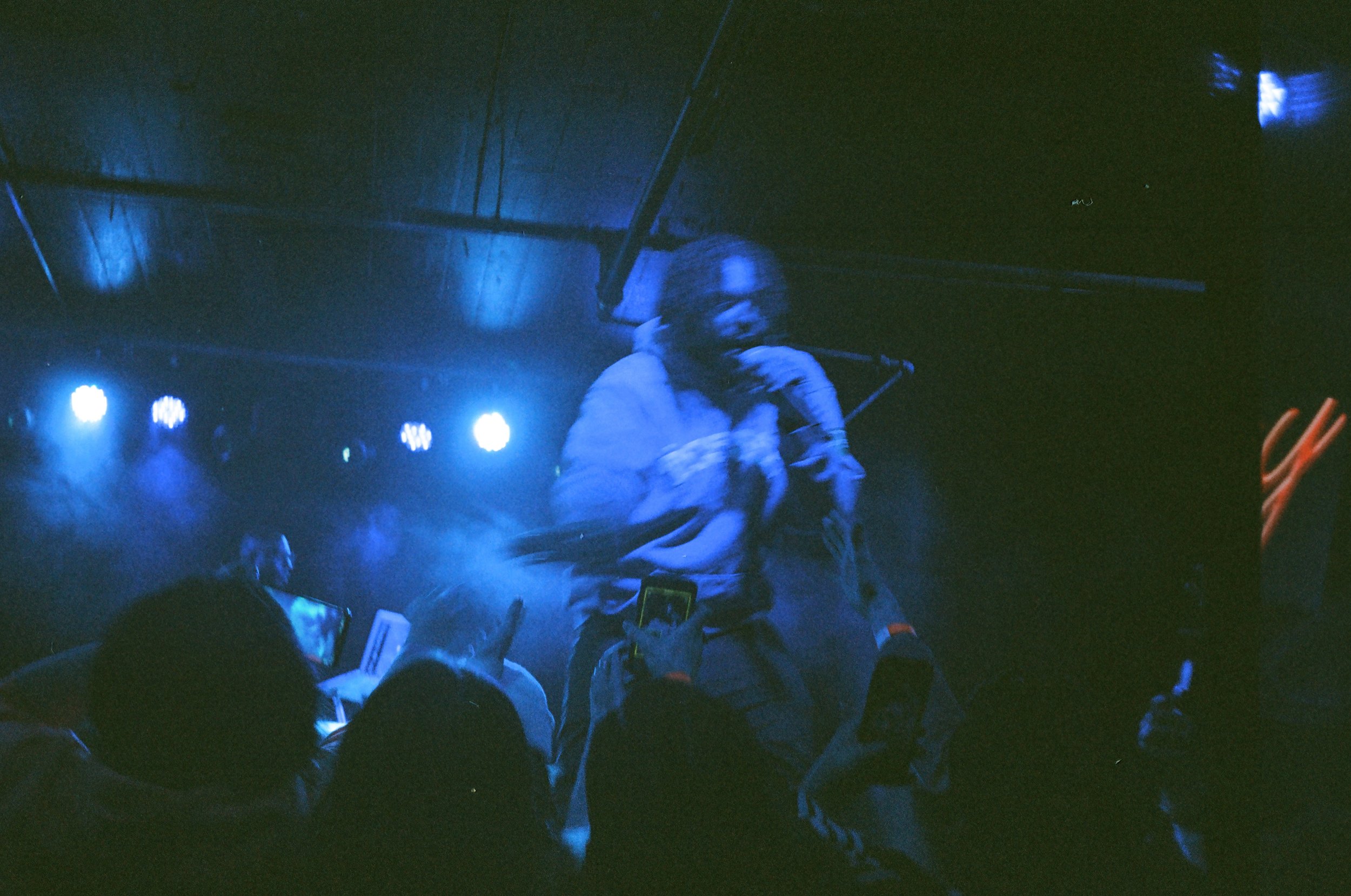 Performer with long hair and glasses singing into a microphone on stage under blue lighting surrounded by audience members taking photos.