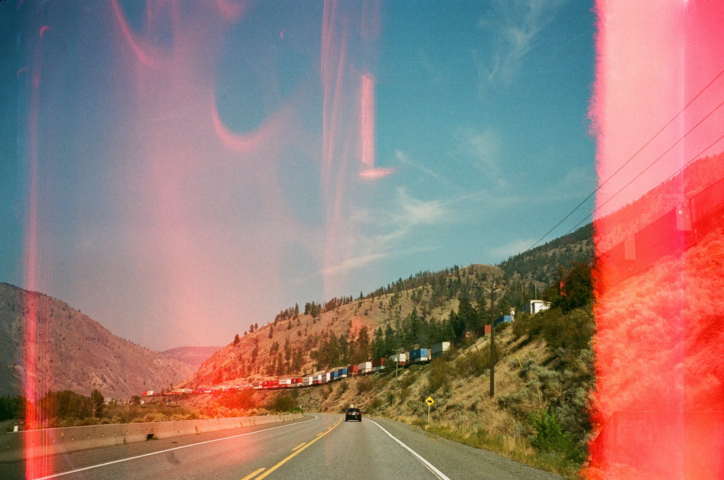 A two-lane road in a mountainous area with a train of cargo containers on the hillside to the right. Clear blue sky with a few clouds and power lines crossing the scene.