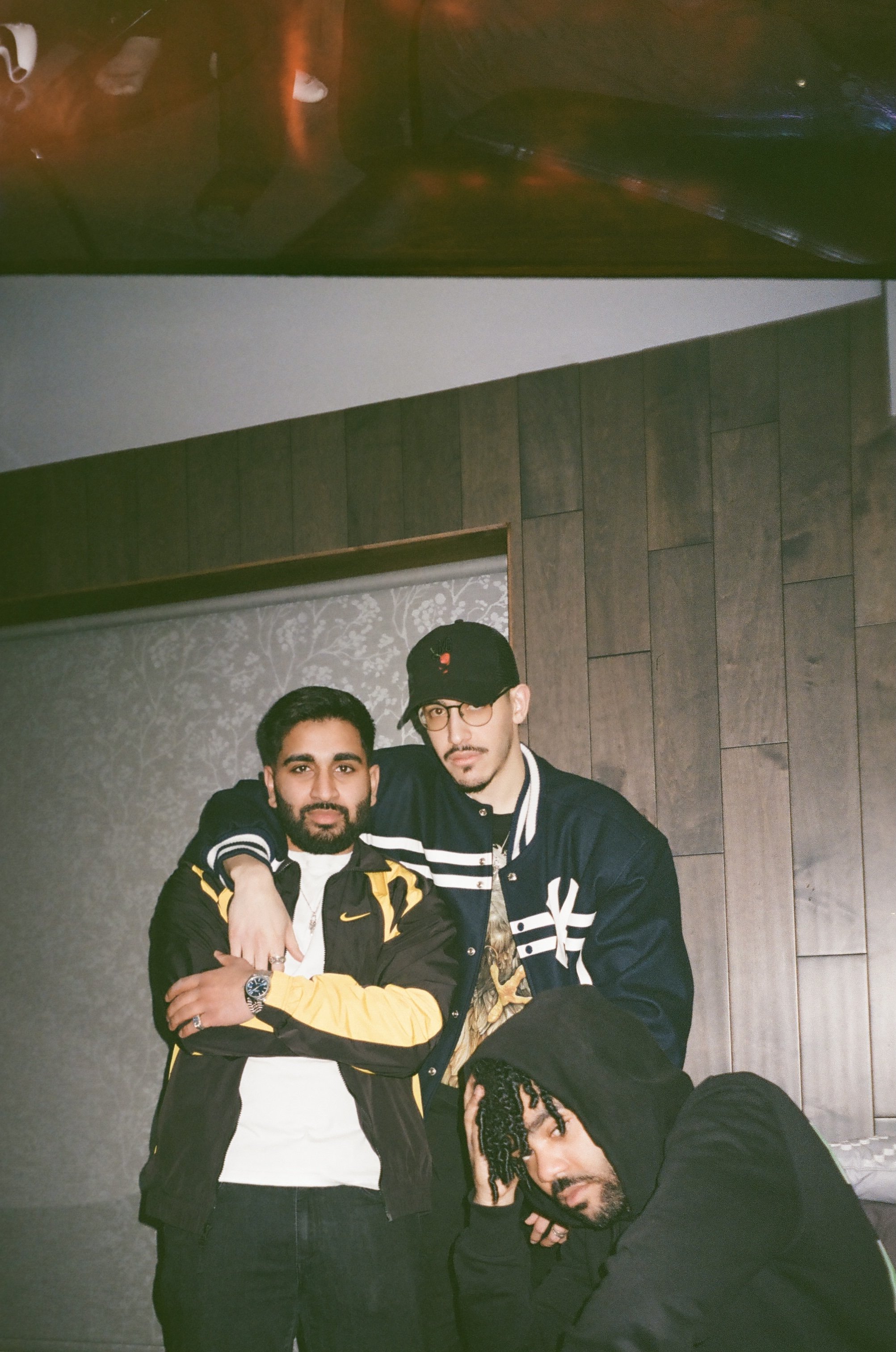 Three young men in casual clothing indoors, two standing and one seated. The two standing men are embracing, one with a yellow and black jacket and the other with a dark baseball cap and glasses. The seated man is wearing a black hoodie and has curly