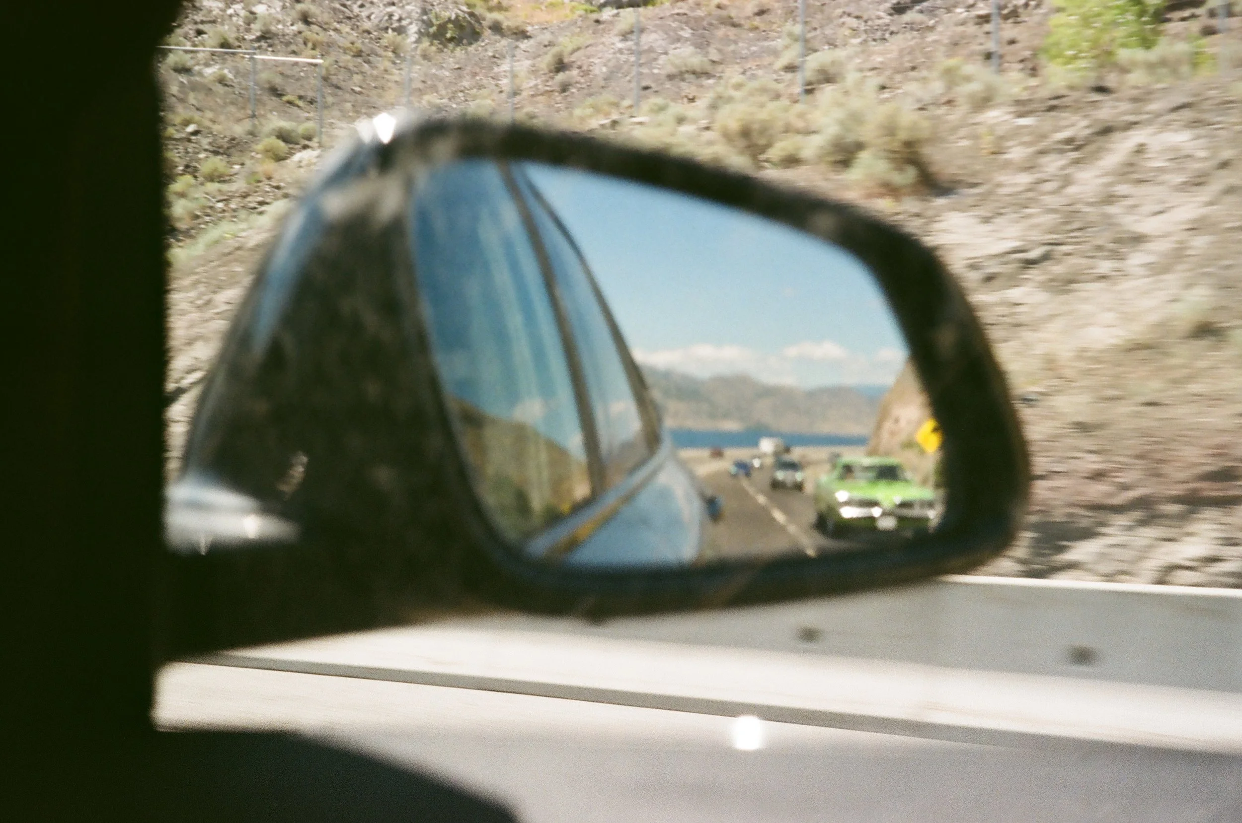View of a highway seen through a car's side mirror, showing multiple vehicles including a green car, with mountains and a partly cloudy sky in the background.