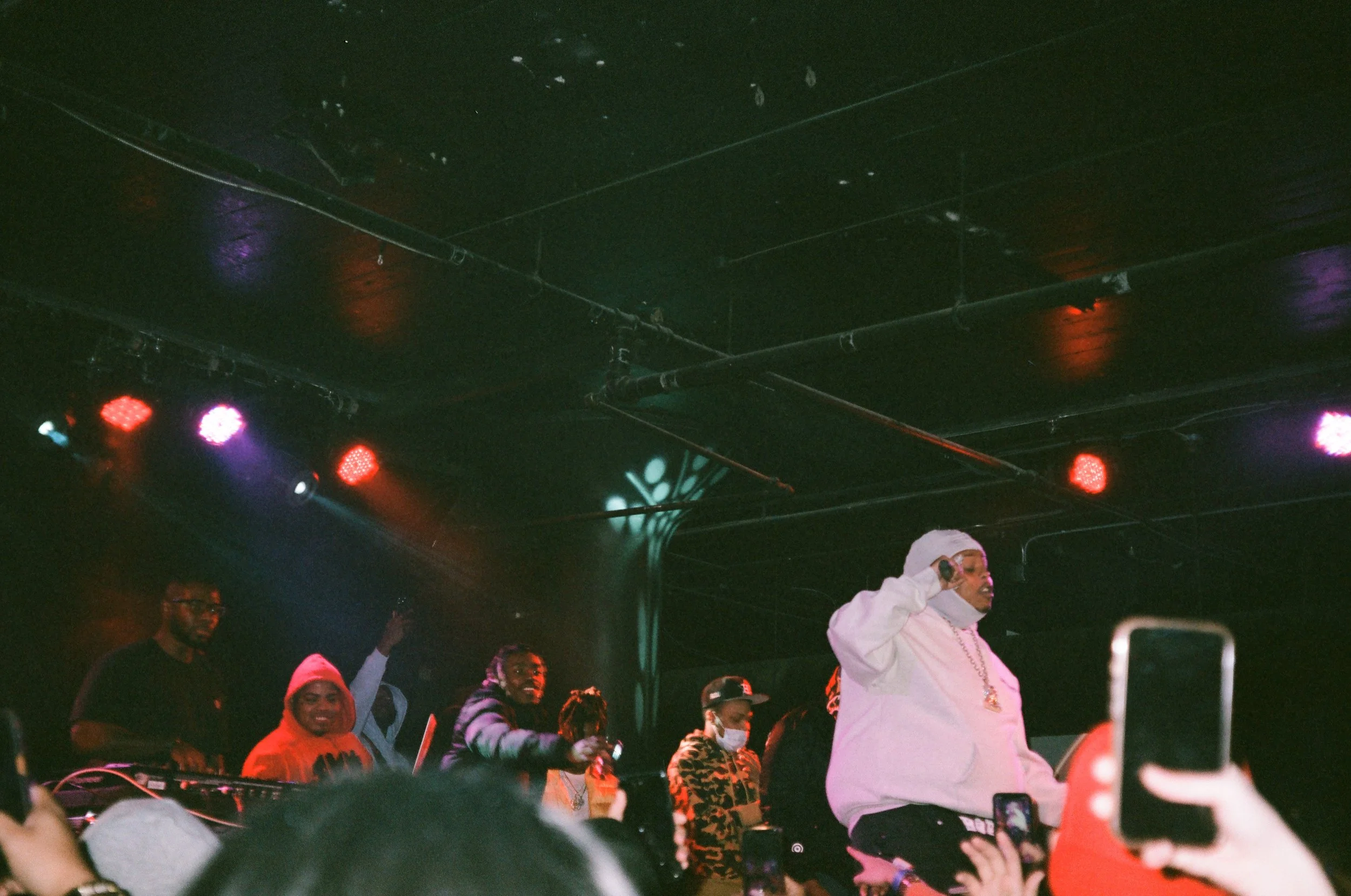 Hip-hop concert with a DJ and rapper on stage, diverse crowd, colorful stage lights, and some audience members taking photos.