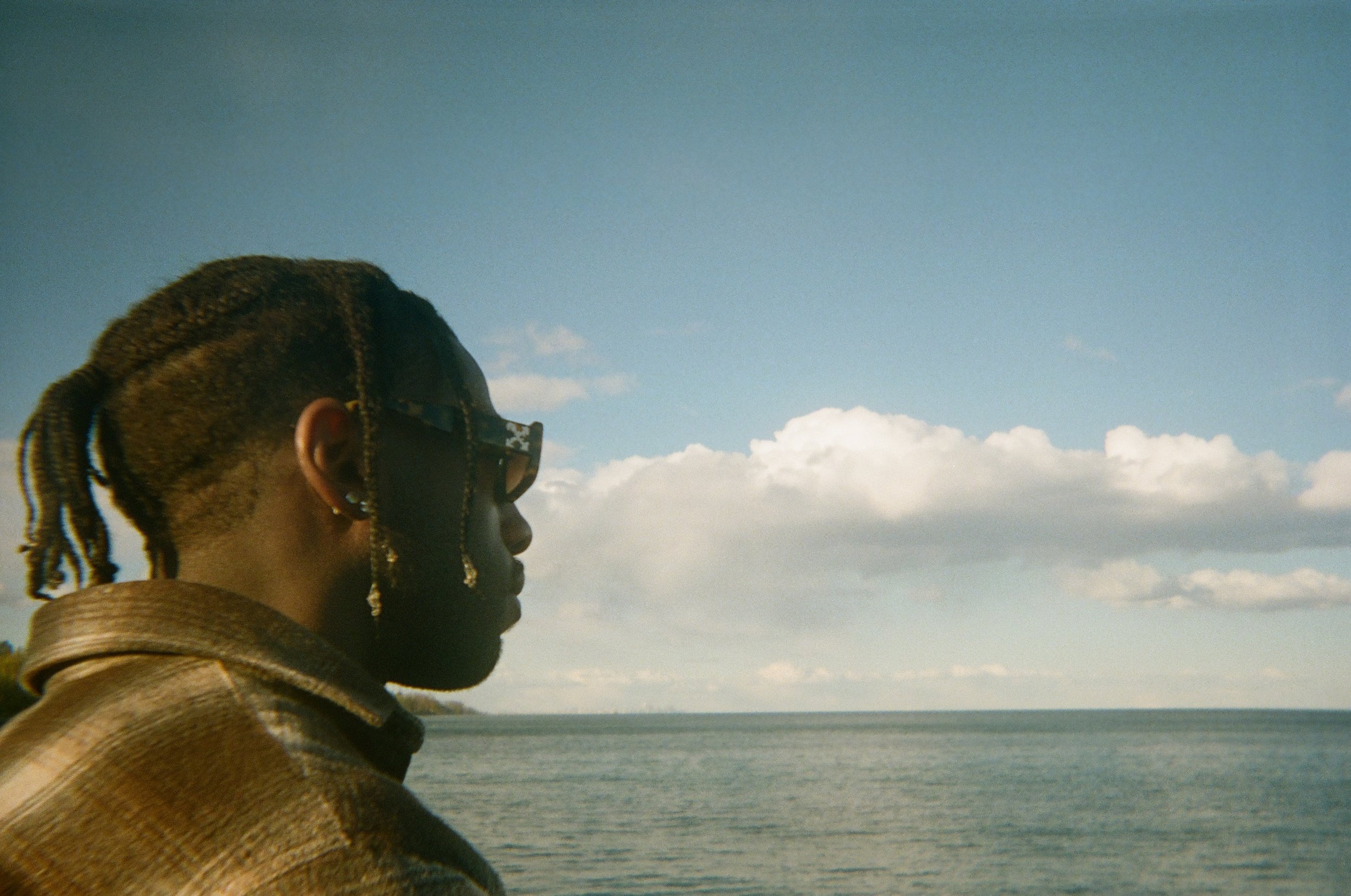 A person with braided hair, wearing sunglasses and a tan jacket, looks out over a body of water with a cloudy sky overhead.