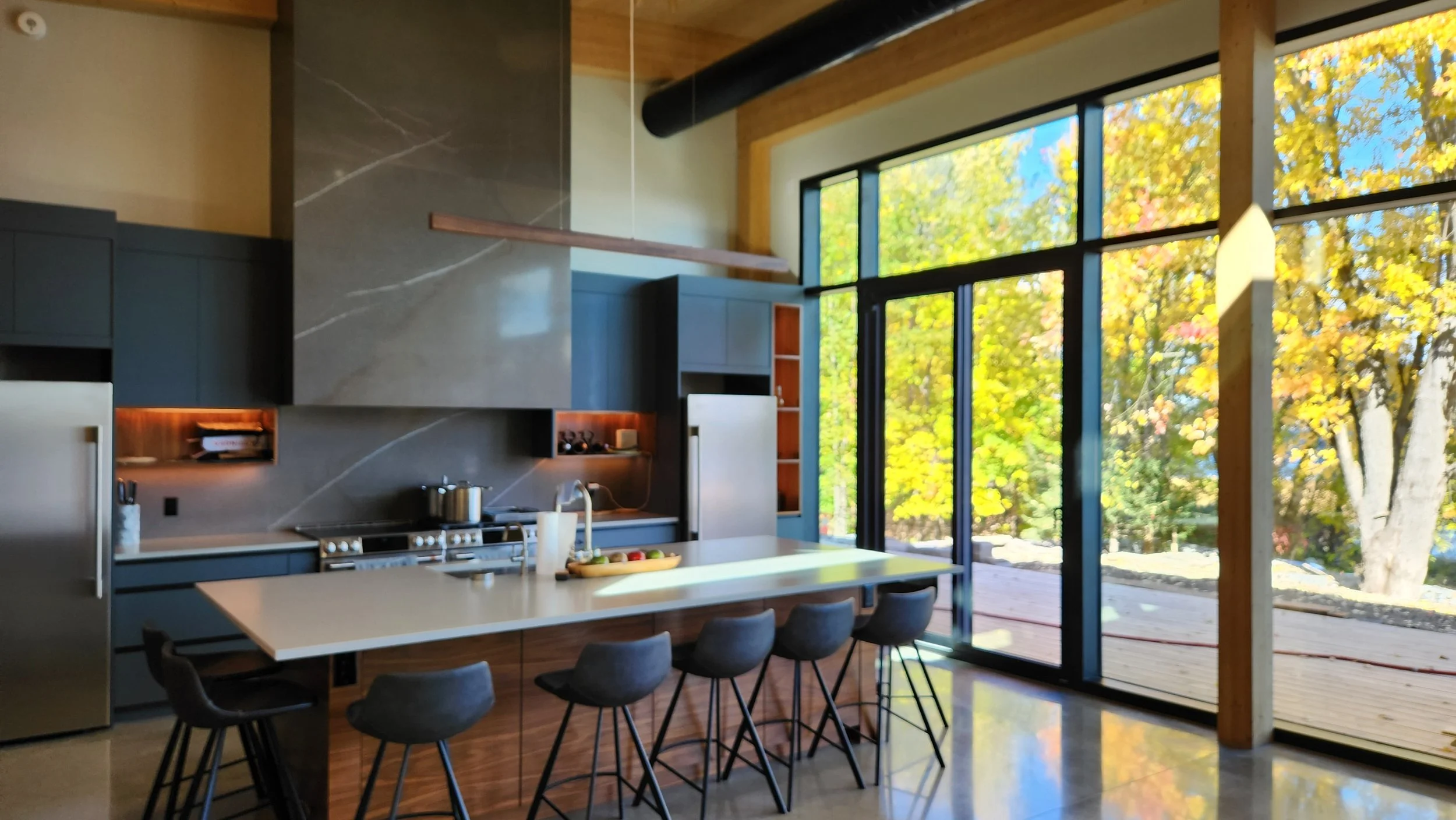 Modern wooden house with large glass window, set against a mountainous landscape with dry grass and low shrubs in the foreground.