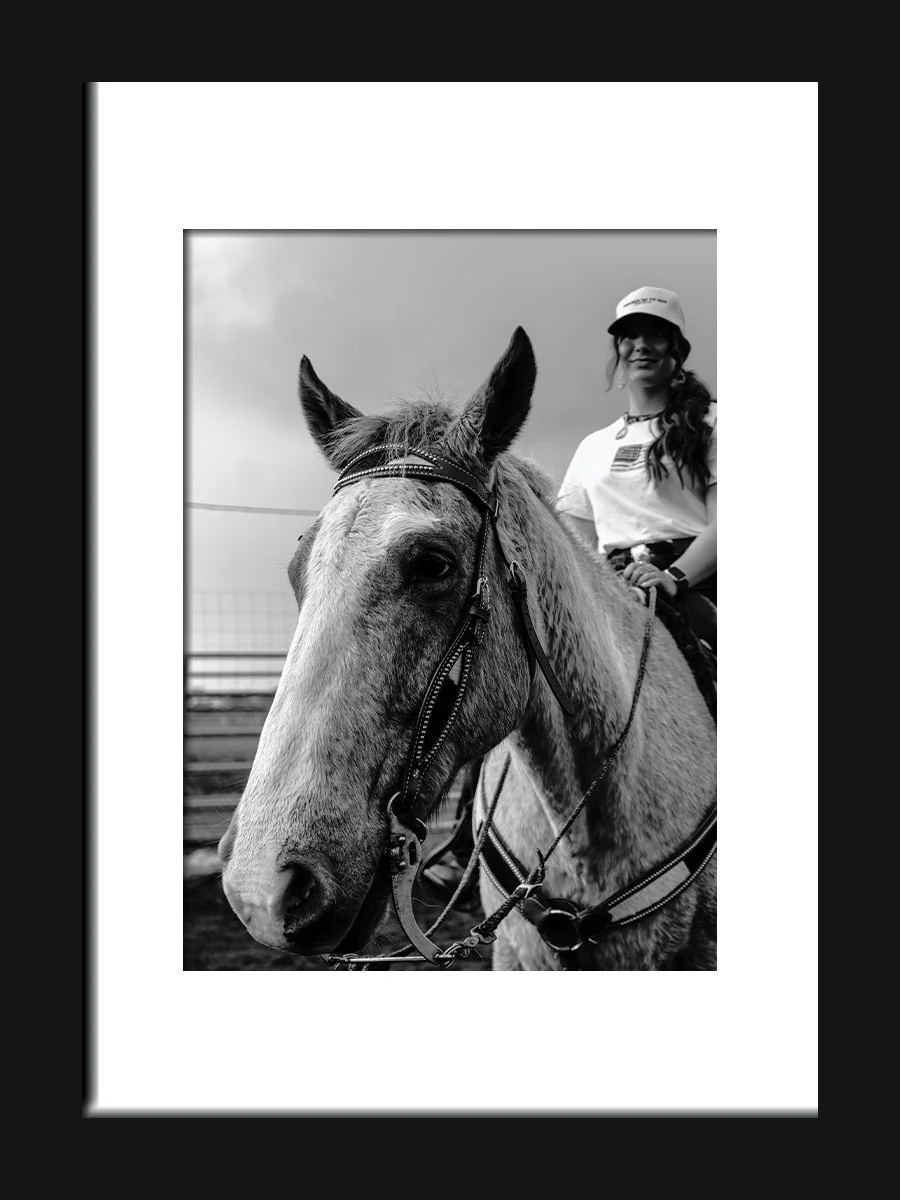 Black and white photo of a woman riding a horse, smiling, wearing a hat and casual clothing, with a cloudy sky in the background.