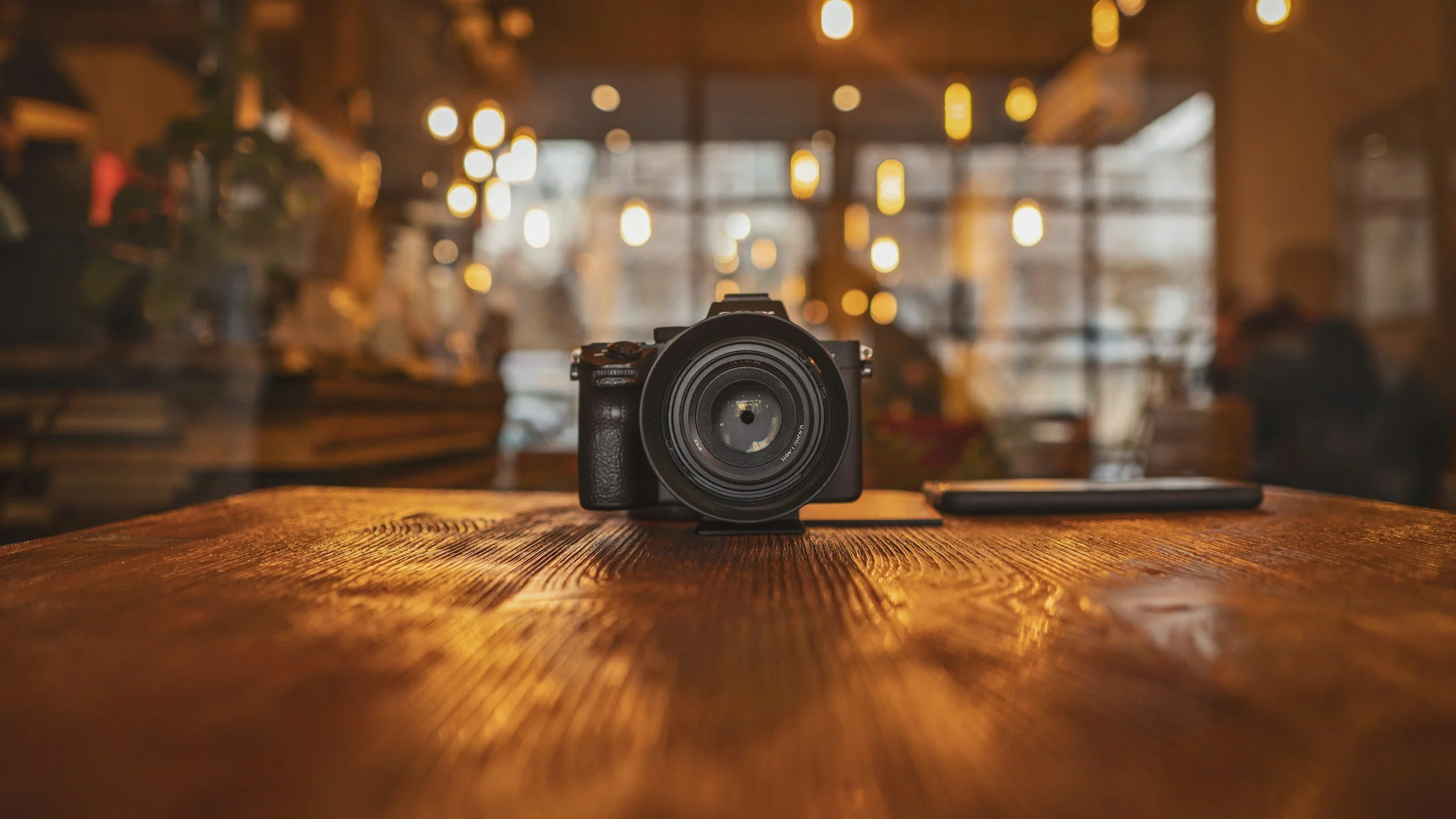 A camera placed on a polished wooden table in a warmly lit cafe or restaurant with blurred lights and background figures.