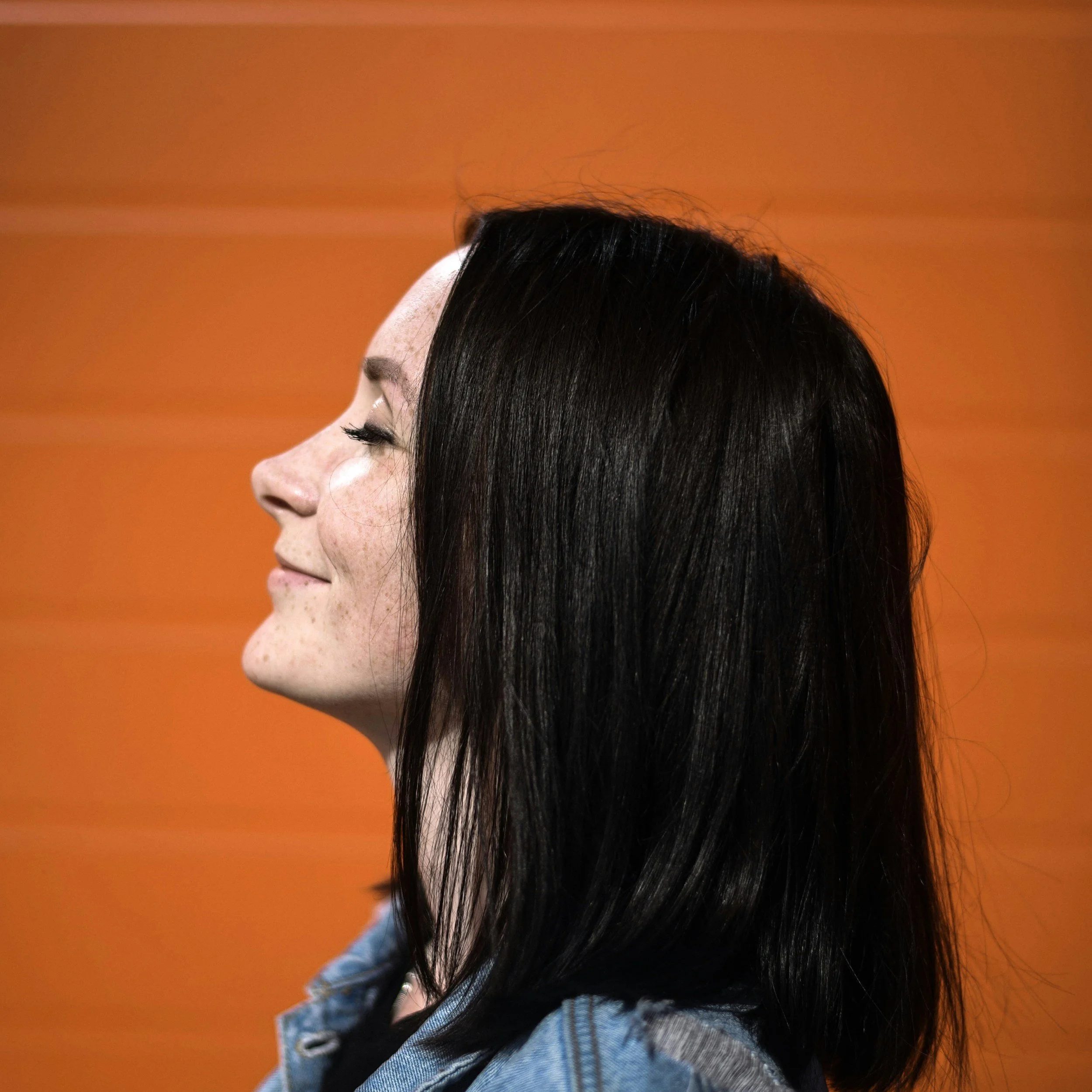 Close-up profile of a woman with black hair and light skin, wearing a blue denim jacket and a black shirt, against an orange background.