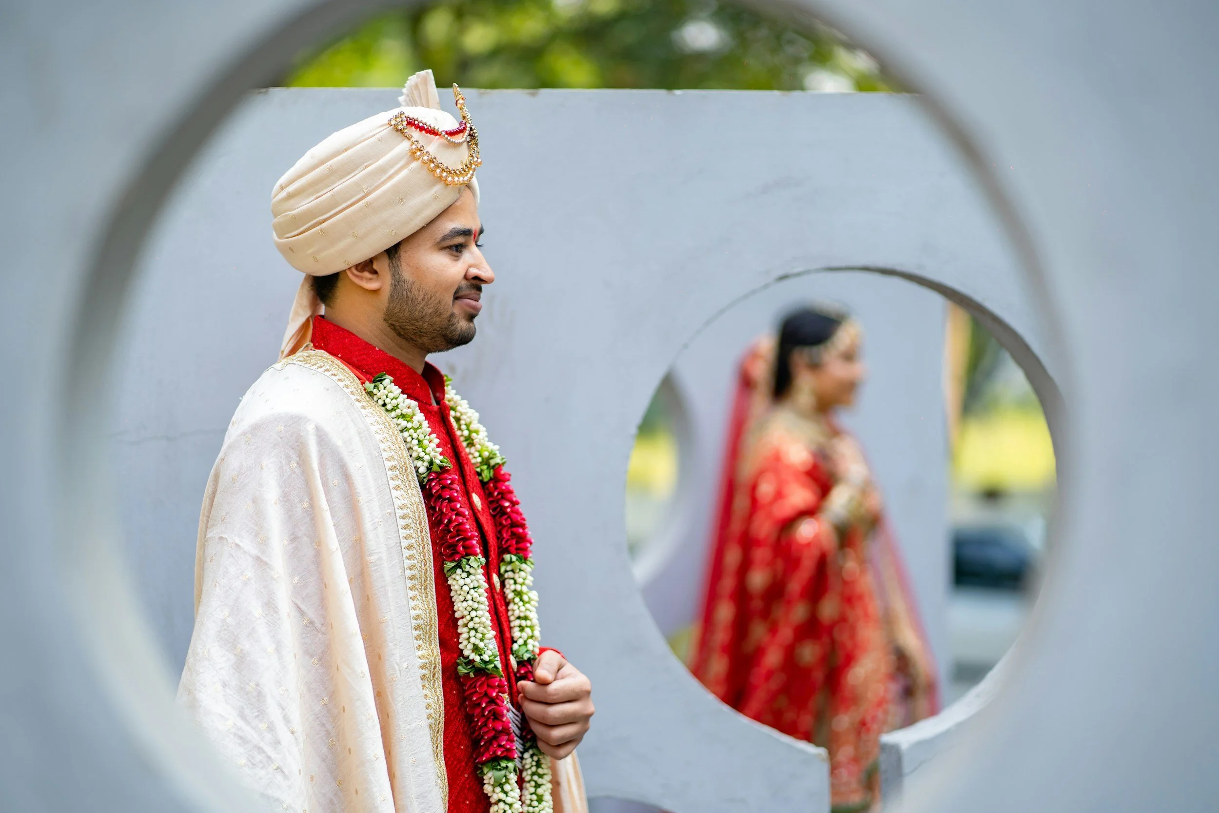 A man dressed in traditional Indian wedding attire, wearing a cream-colored turban adorned with jewelry and a garland, is seen through a heart-shaped opening, with a woman in similar attire blurred in the background.