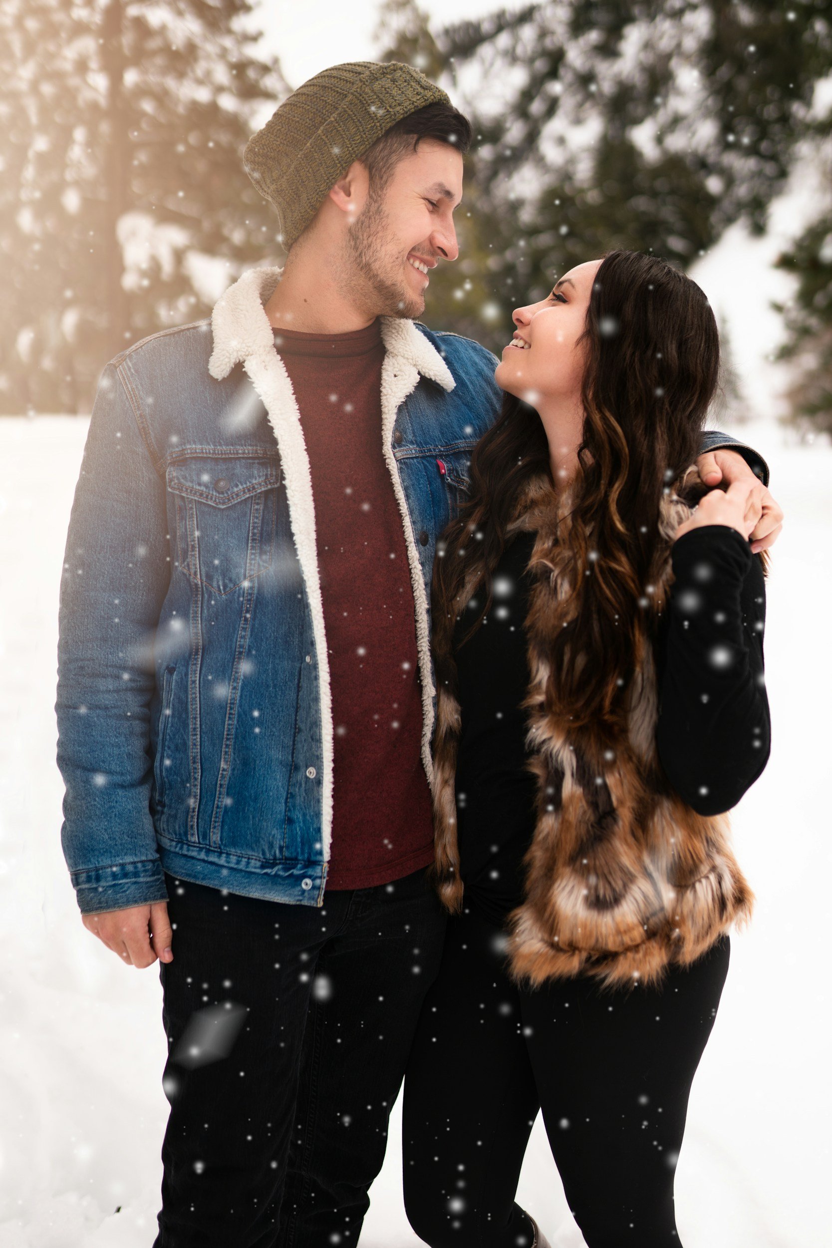 A young man and woman standing close together outside in a snowy winter setting, smiling and looking into each other's eyes, with snow falling around them.
