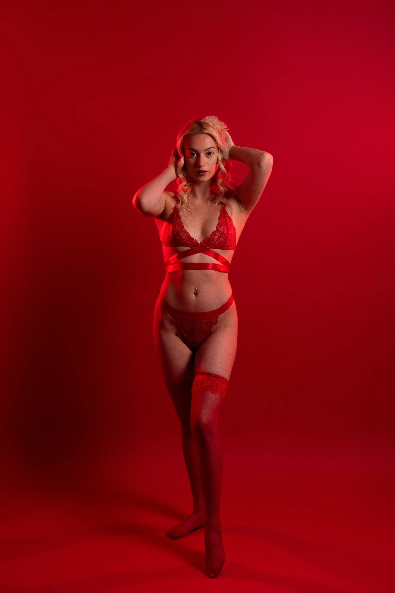 A woman with blonde hair in loose curls, wearing red lace lingerie and thigh-high stockings, standing against a red backdrop, posing with her hands in her hair.
