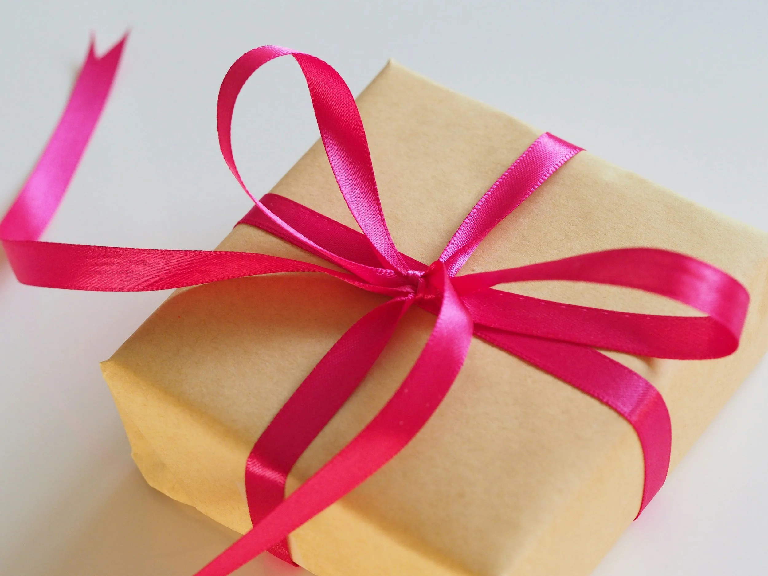 A beige gift box wrapped with a pink satin ribbon tied in a bow, with the ribbon streaming off the side.