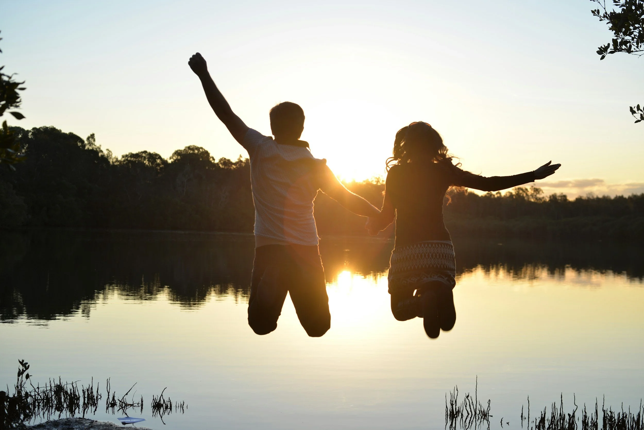 Silhouettes of a man and woman holding hands, with one hand raised and the other extended, standing by a calm body of water at sunset.