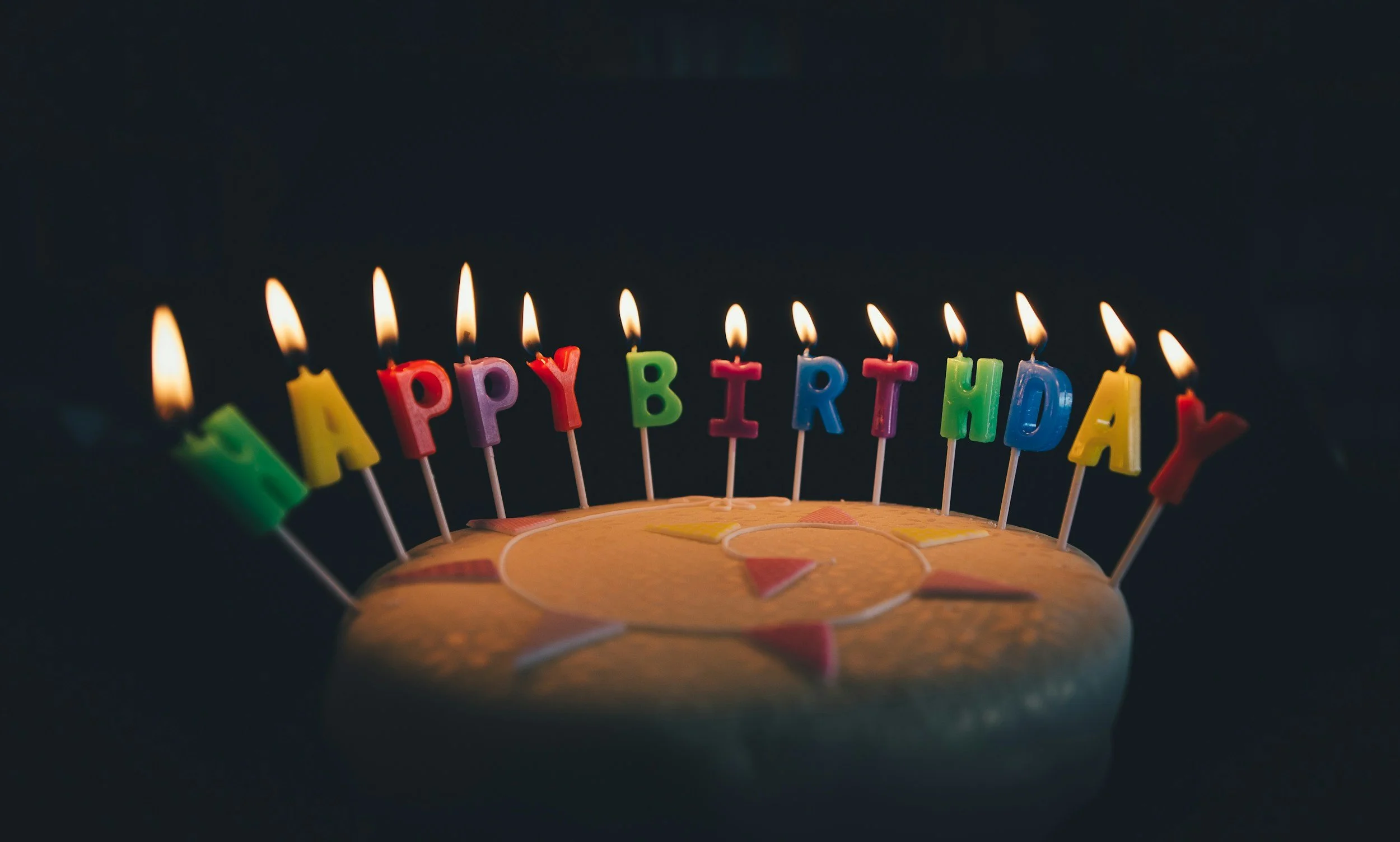 A birthday cake with colorful lit candles spelling 'HAPPY BIRTHDAY' on a dark background.