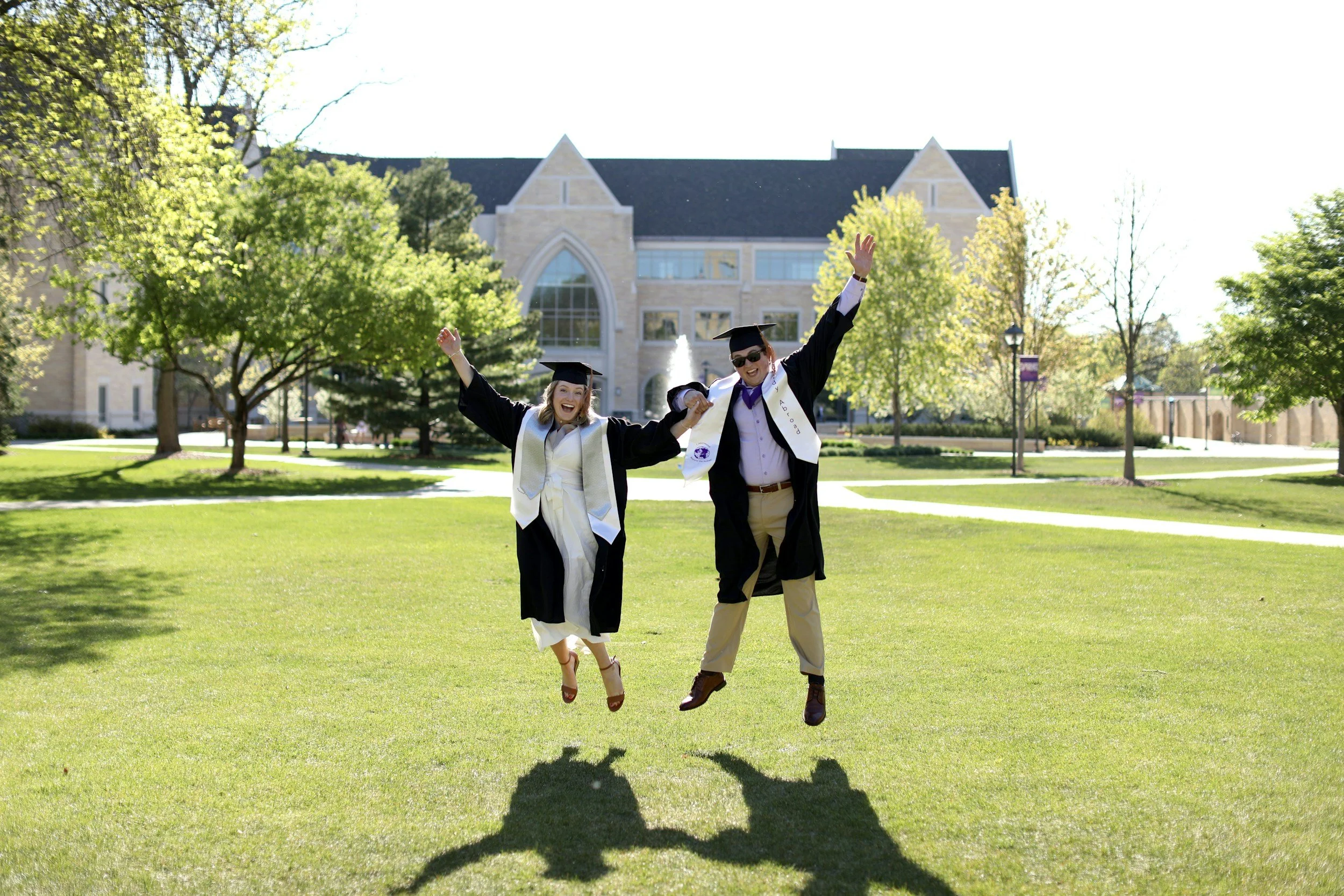 Two graduates in caps and gowns jumping in the air on a green lawn in front of a large stone building with arched windows, surrounded by trees with green leaves, on a sunny day.