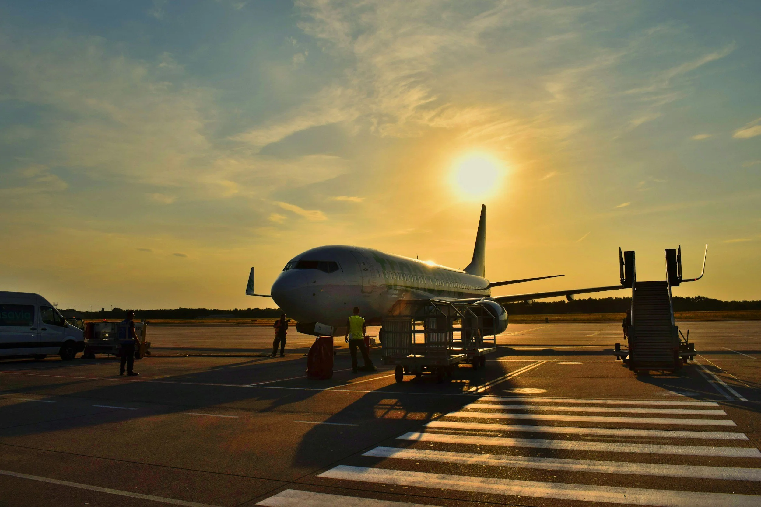 Airplane parked at the gate during sunset with airport ground crew and luggage carts nearby.