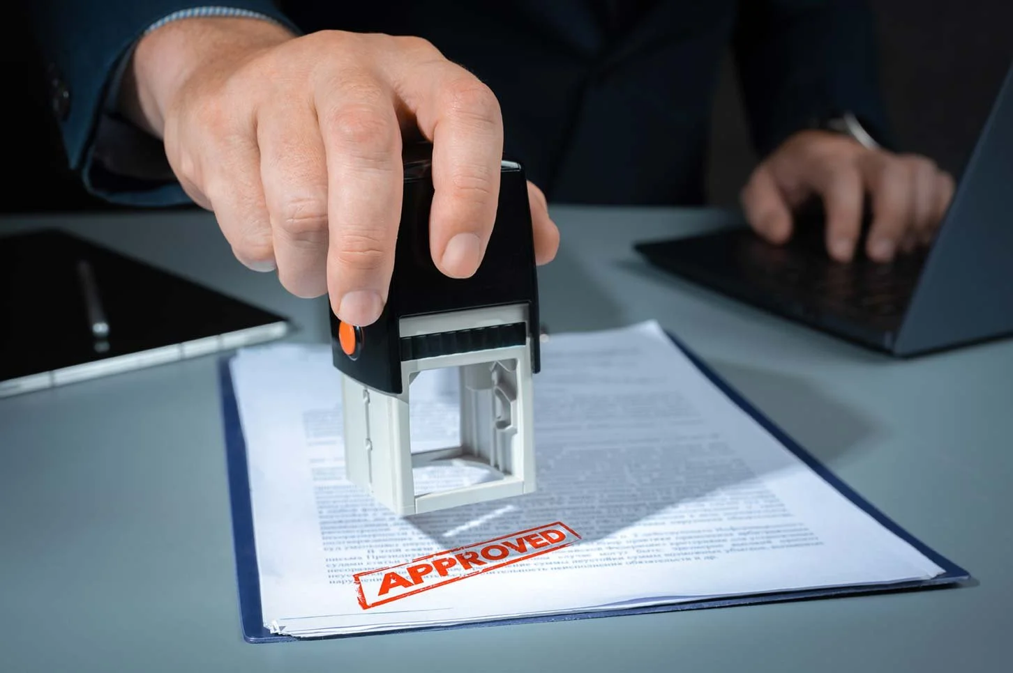 A person using a stamp to mark an approval on a document on a desk with a laptop and pen nearby.