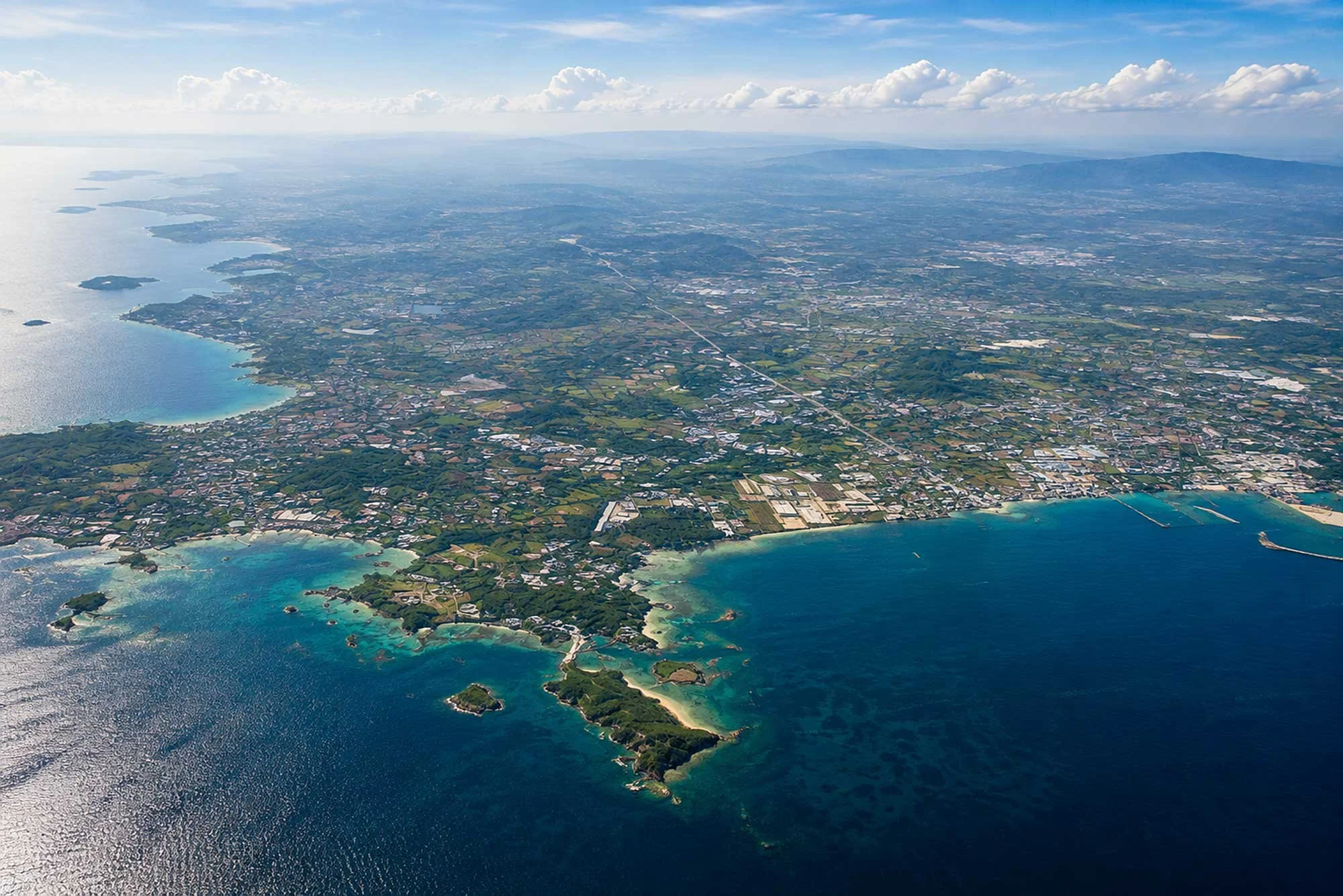 Aerial view of a coastline with turquoise waters, islands, and a mix of greenery and urban areas extending inland under a partly cloudy sky.