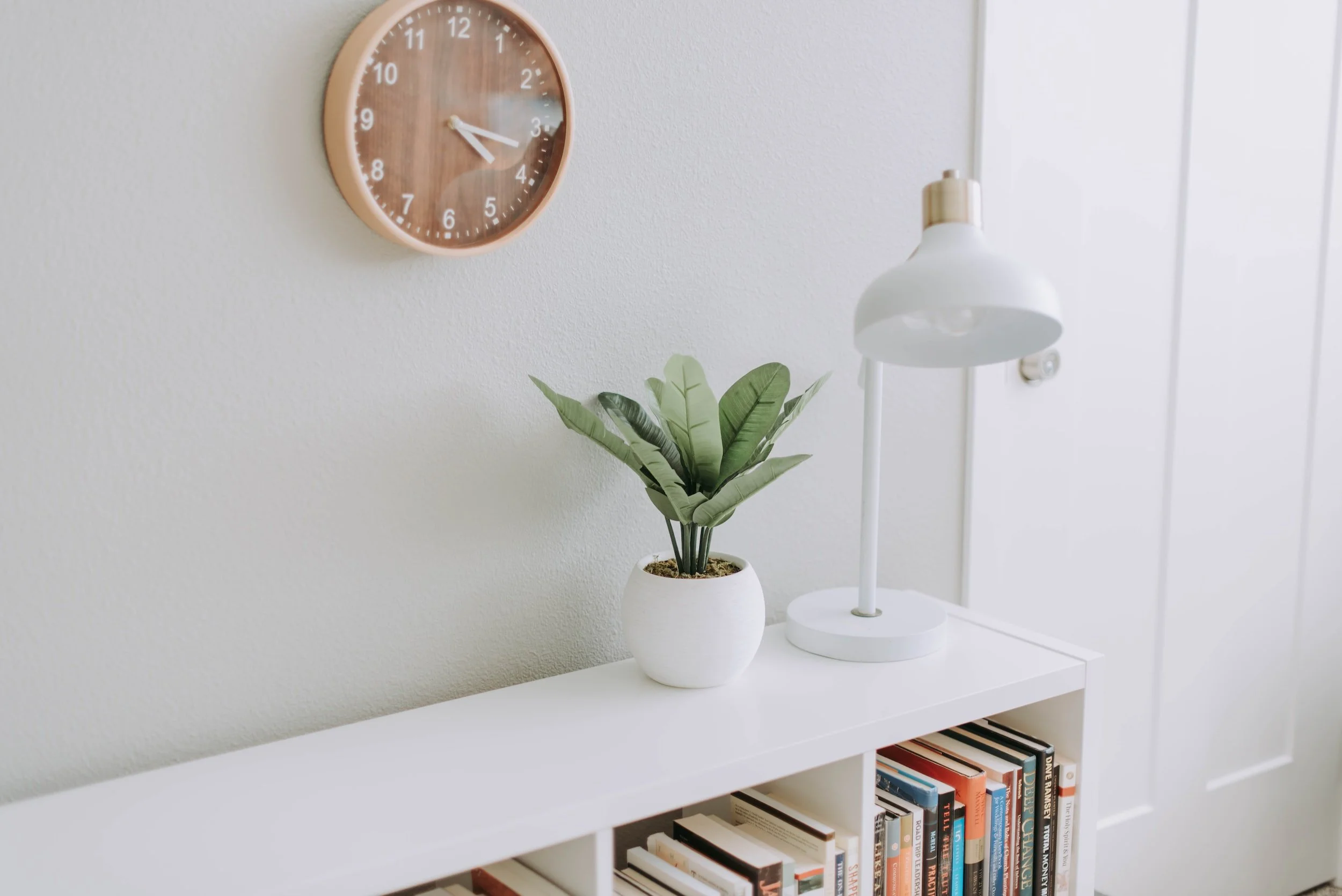 A white bookshelf with a potted green plant and white desk lamp on top, against a white wall with a round wooden wall clock showing 4:44.