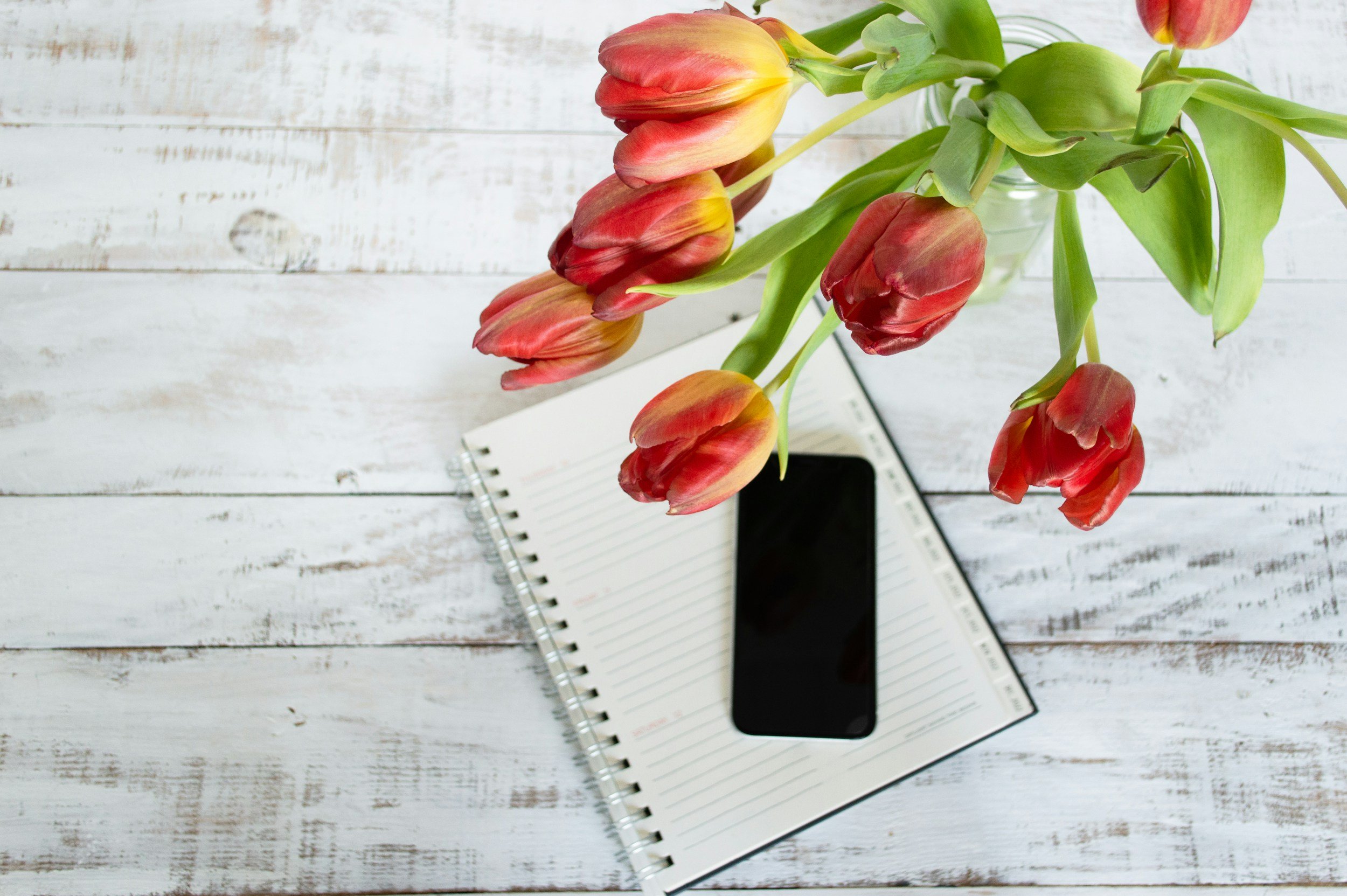 A bouquet of red and yellow tulips in a glass vase placed on an open notebook with lined pages, with a smartphone resting on top of the notebook. The background is a white wooden surface.
