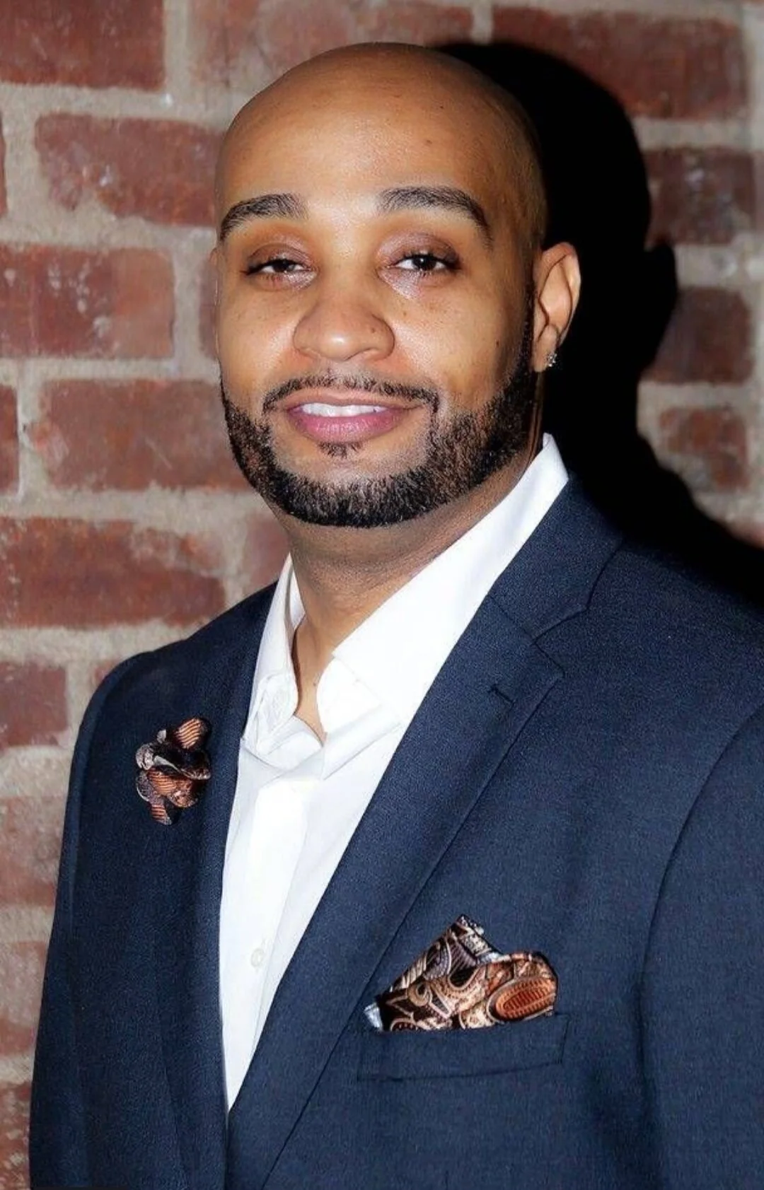 A man with a shaved head and beard wearing a dark suit, white shirt, and patterned pocket square, standing against a brick wall.
