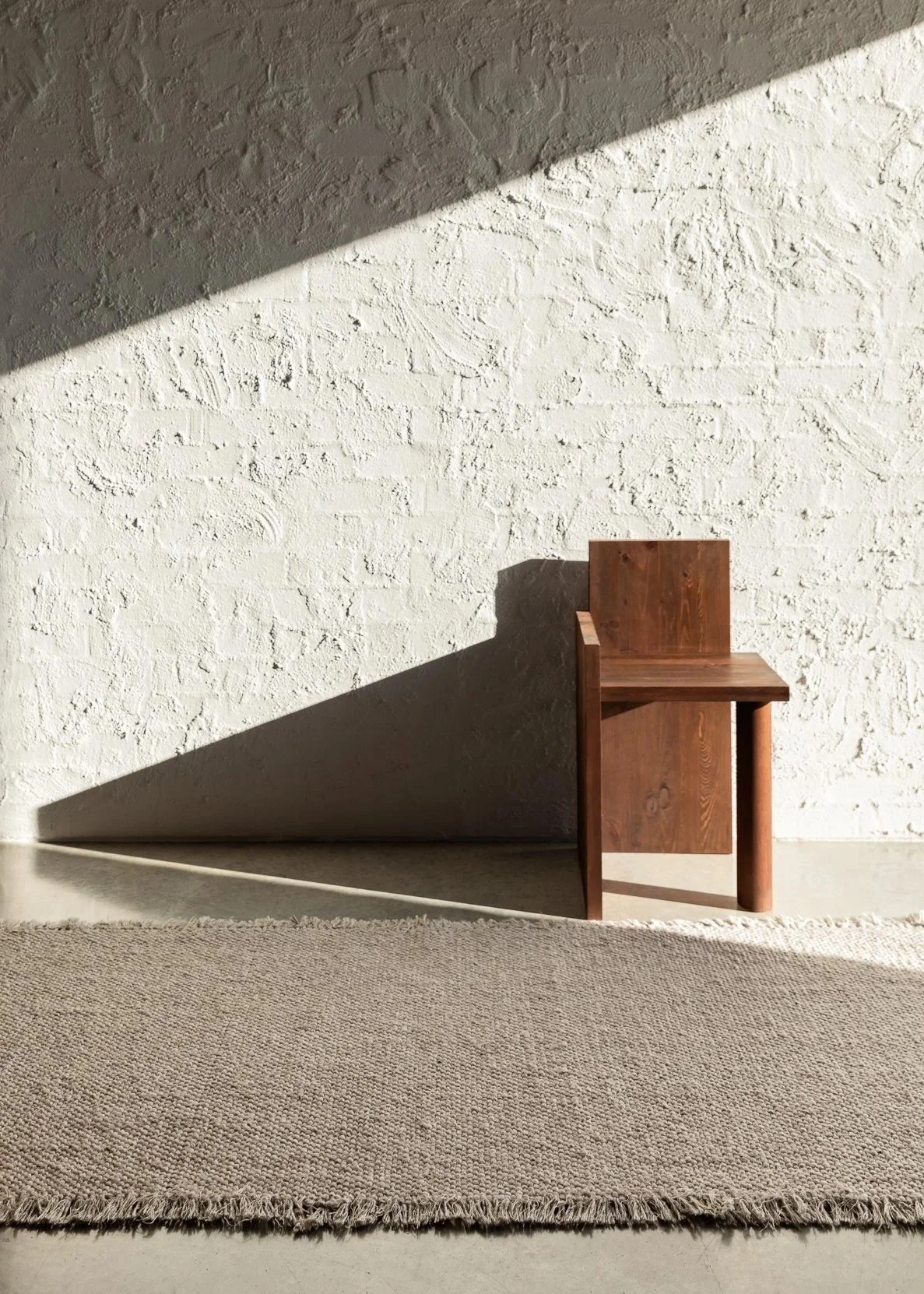 Modern timber chair sitting in sunlight entrance space with strong shadows against a white plastered wall and floor rug.