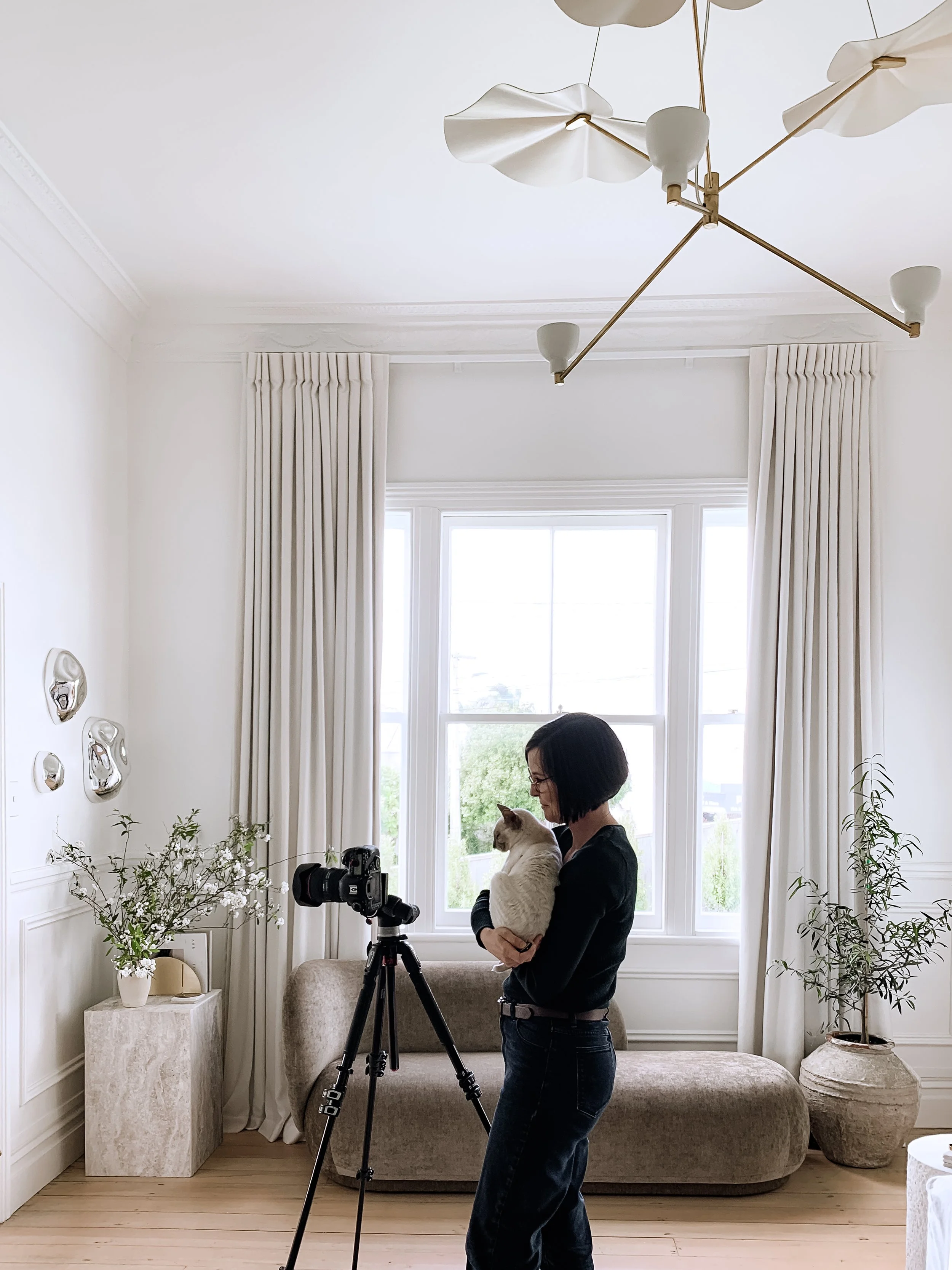 Woman standing in a bright living room holding a cat while setting up a camera on a tripod.