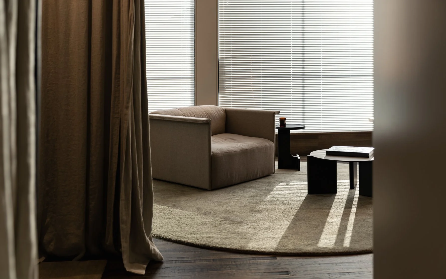 A cozy changing room waiting area with a beige sofa, two black side tables, a candle, books, and large window blinds.