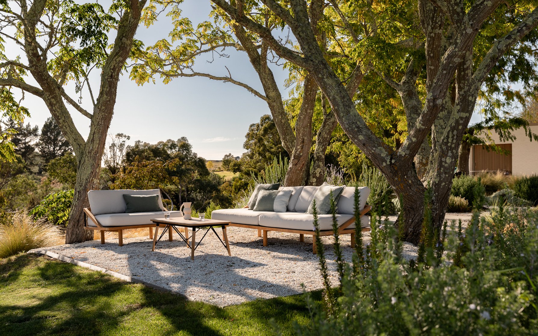 Outdoor seating area with white cushioned sofas and a small wooden coffee table on a gravel surface, shaded by large trees with green foliage, overlooking a scenic landscape with trees and fields.