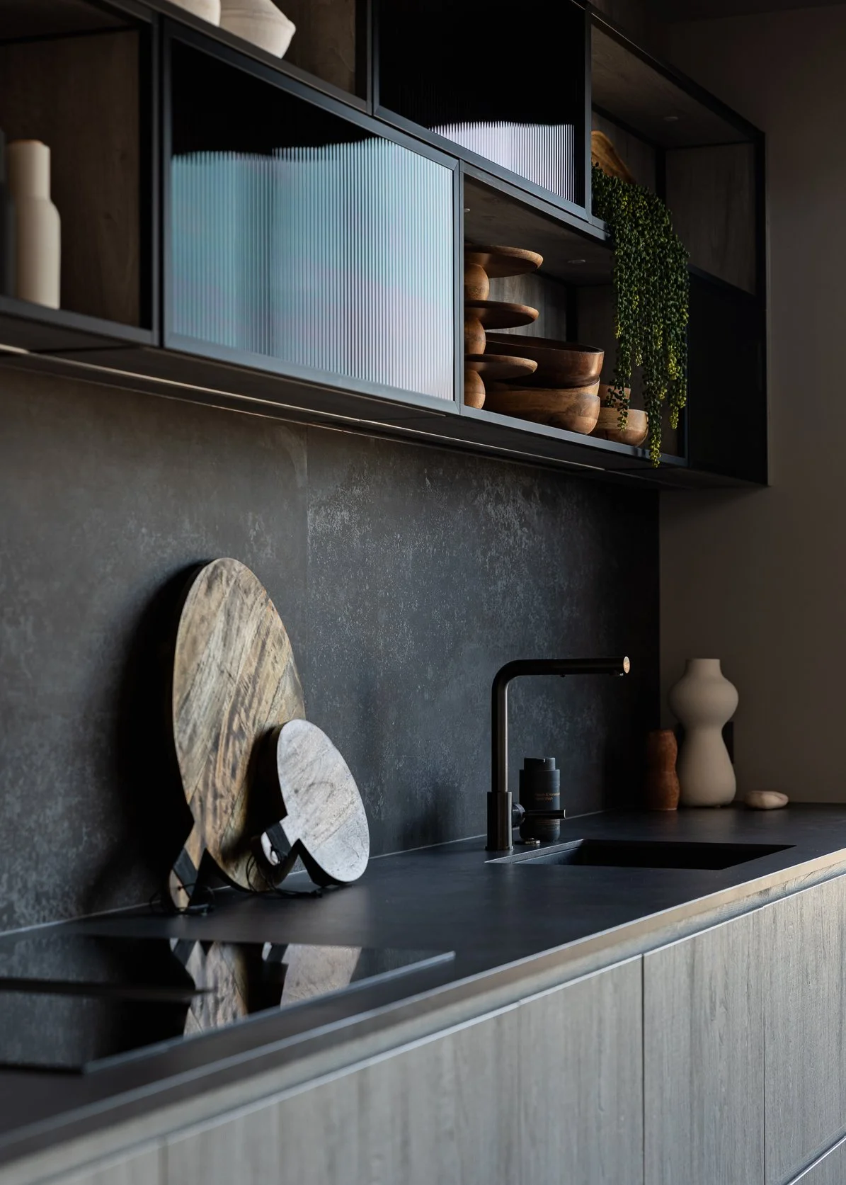 Modern kitchen with dark countertop, decorative wooden bowls, cutting boards, vases, and a black faucet, with dark upper cabinets and a textured wall.
