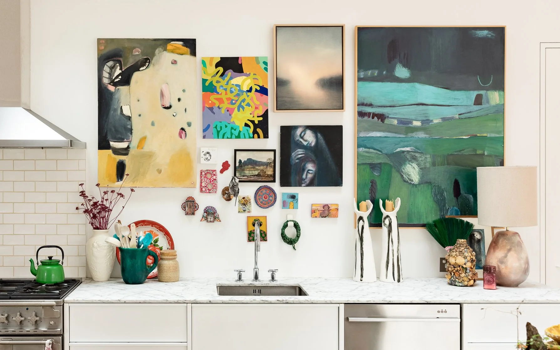 Decorative kitchen wall with various colorful paintings and small decorative items above a white marble countertop with a sink, green teapot, vases, and a lamp.