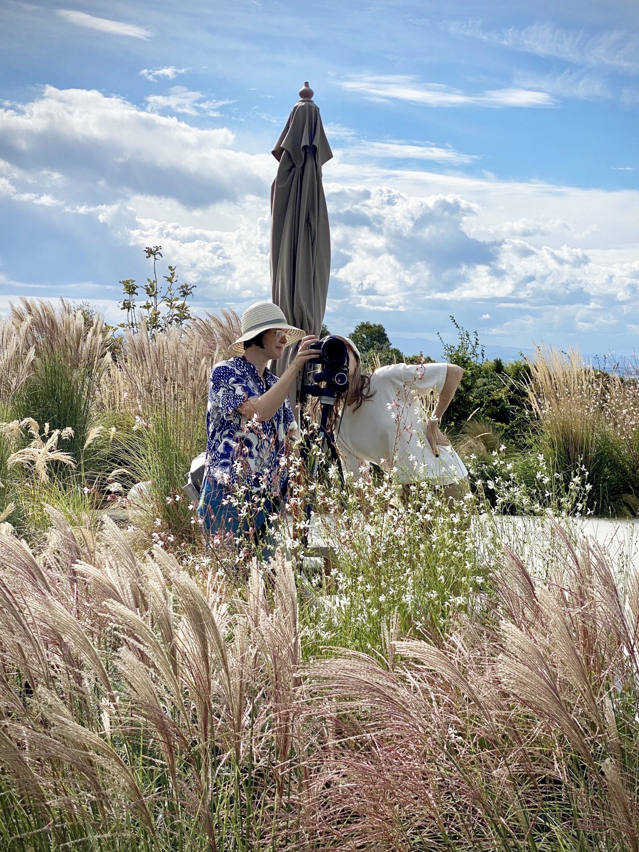 Two women are taking a photo together outdoors in a field of tall grasses and wildflowers, with a cloudy sky in the background.