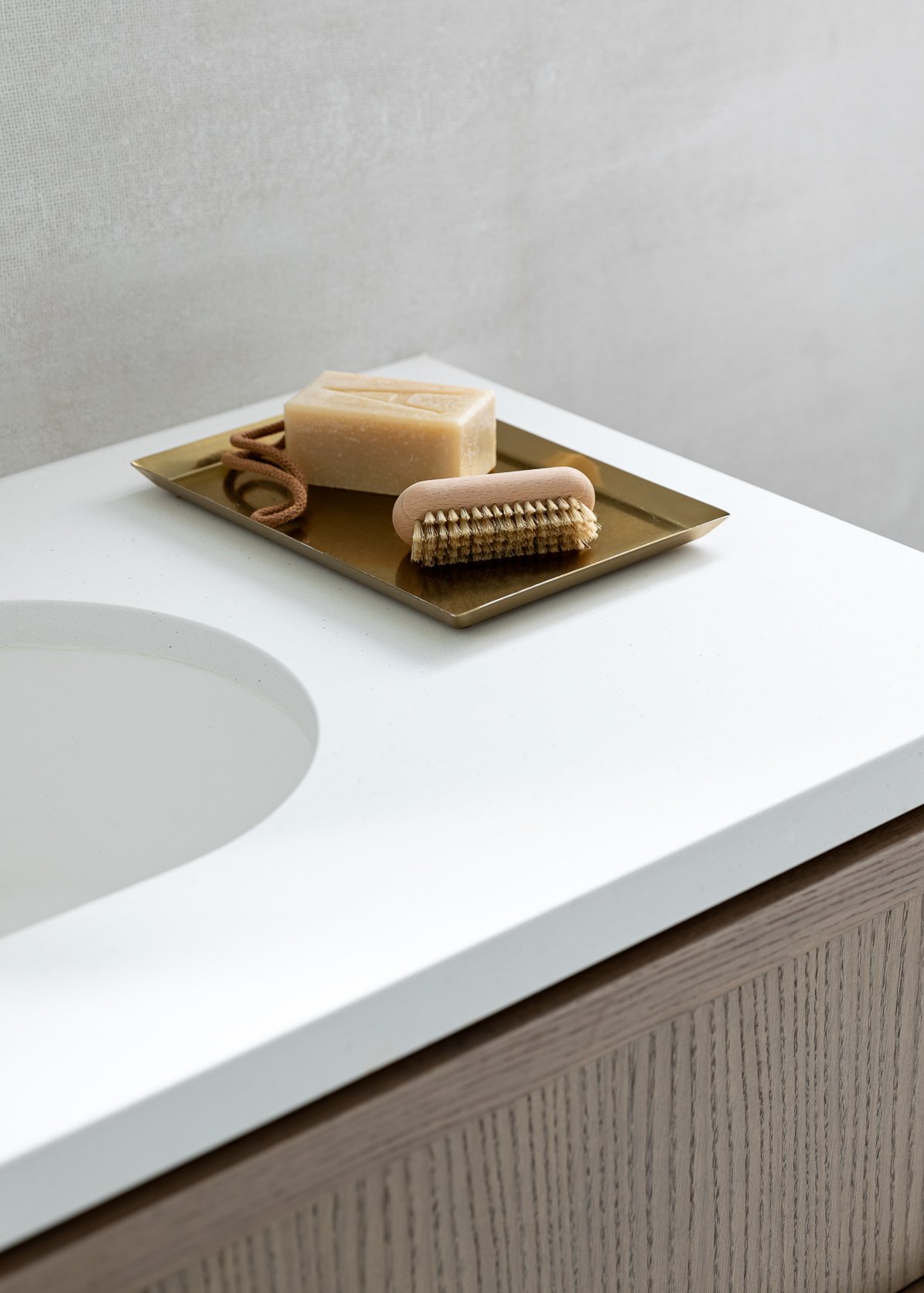 Close-up of a soap bar, a brush, and a string on a gold tray placed on a white bathroom counter near a sink.