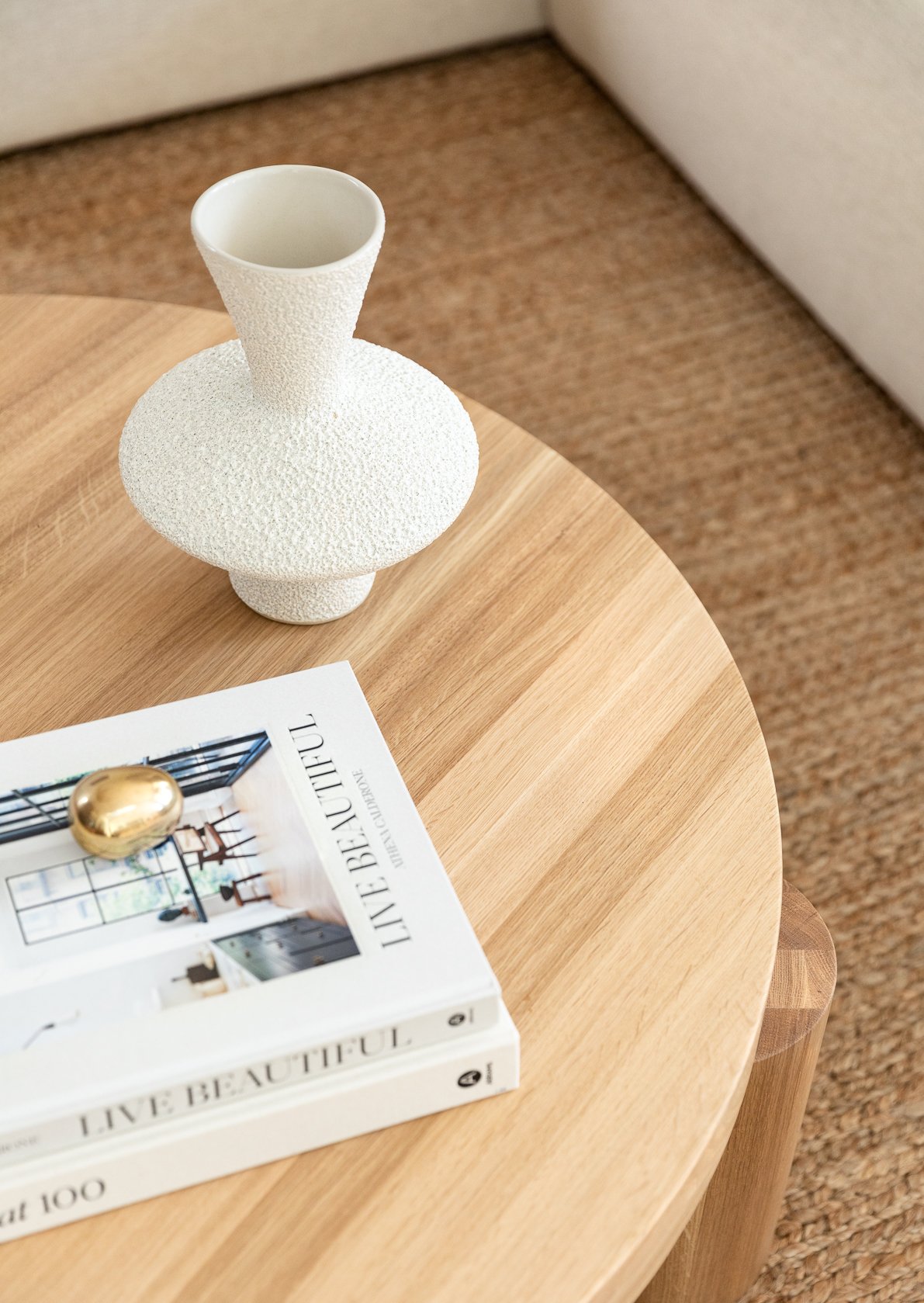A white textured vase and two stacked books titled 'Live Beautiful' are on a round wooden table, with a golden decorative object on top of the books, and a beige carpet and part of a white chair in the background.