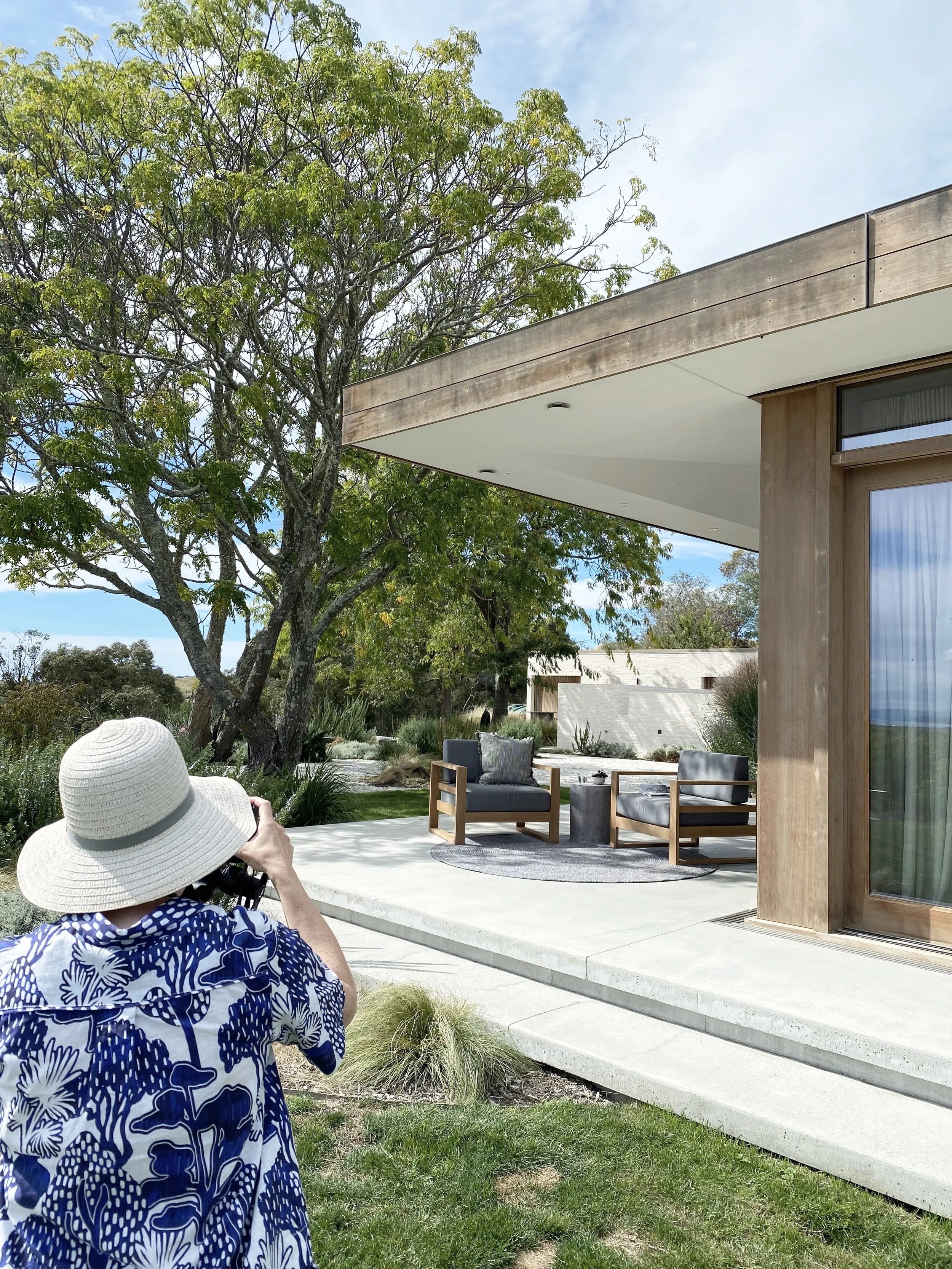 A woman wearing a white sunhat and a blue and white patterned shirt taking a photo of an outdoor patio area with modern chairs, a small table, and a round rug, surrounded by greenery and trees under a partly cloudy sky.