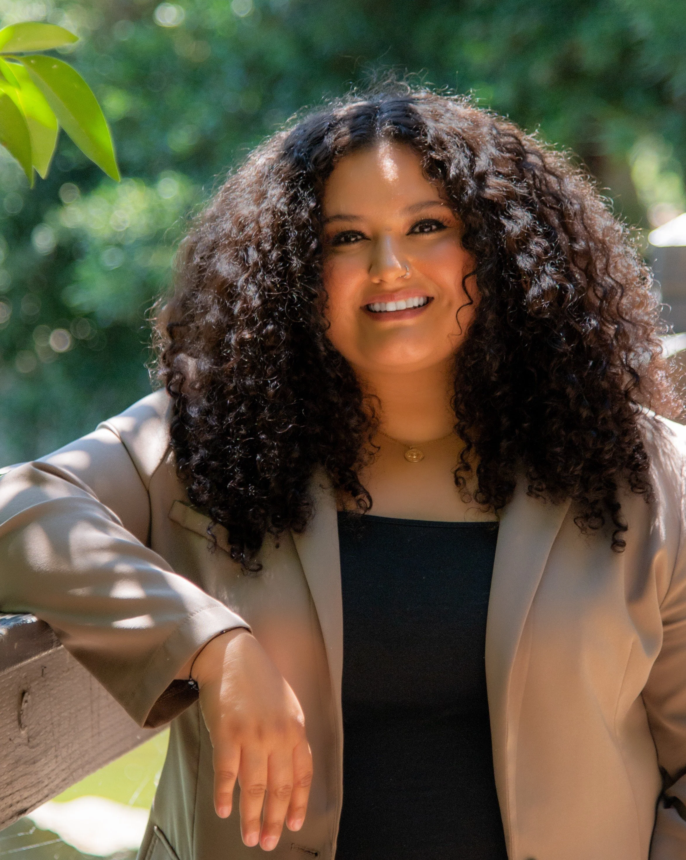 A woman with curly hair smiling outdoors, wearing a beige blazer over a black top, with a blurred green leafy background.