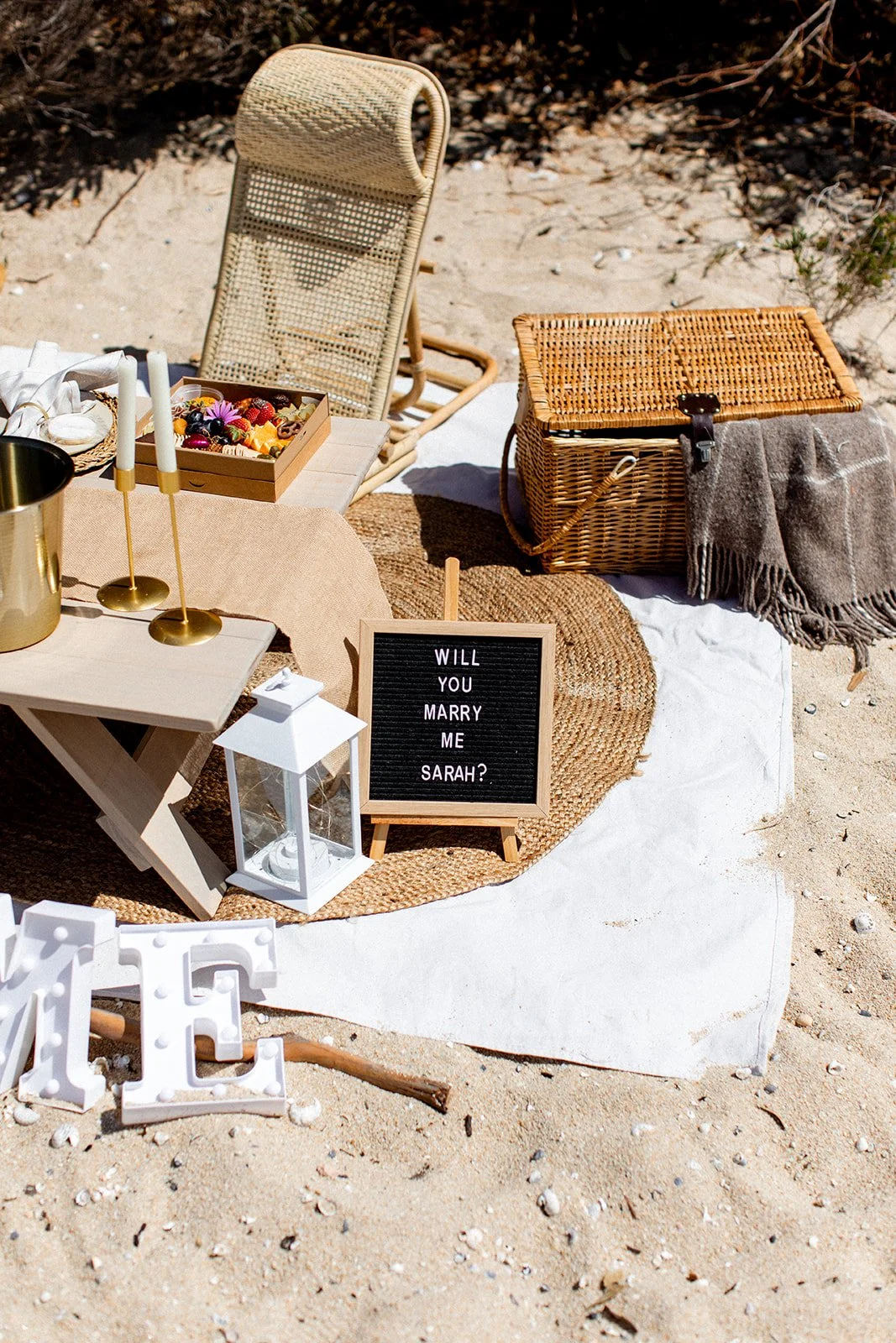 Beach proposal setup with a small table, candles, flowers, a sign reading "Will You Marry Me Sarah?", a lantern, a wicker picnic basket, and a blanket on the sand.