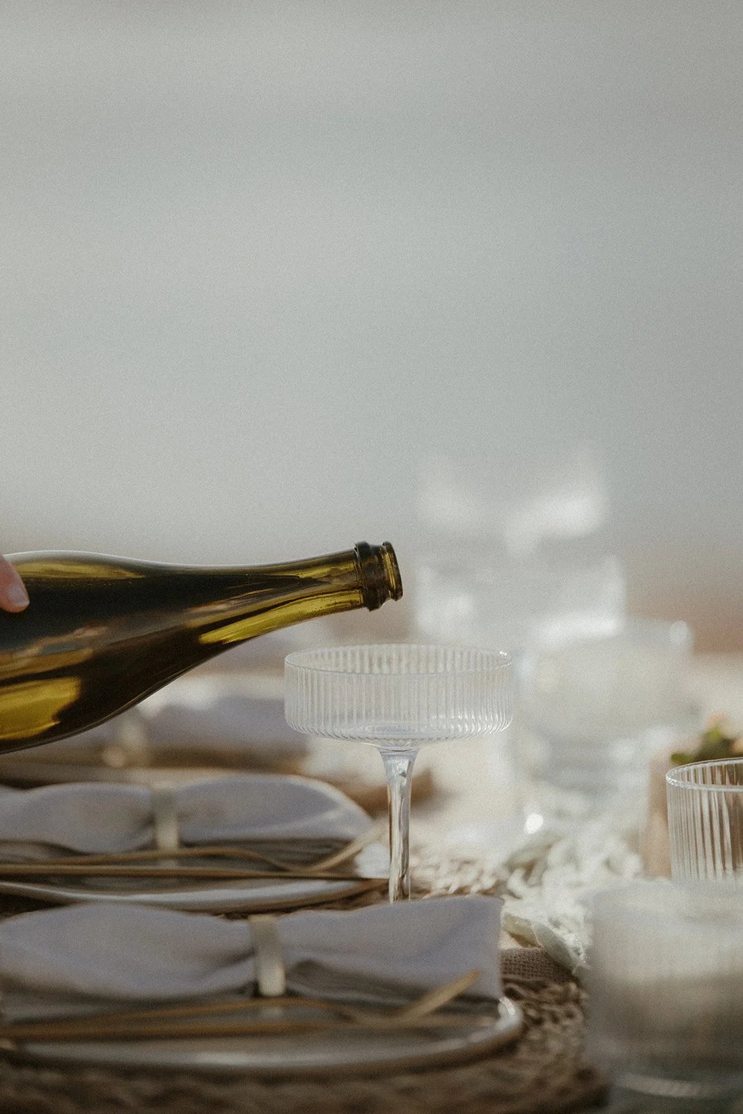 A person is pouring a dark liquid, likely wine or oil, from a brown glass bottle into a clear glass dish on a table set with plates, glasses, and napkins.