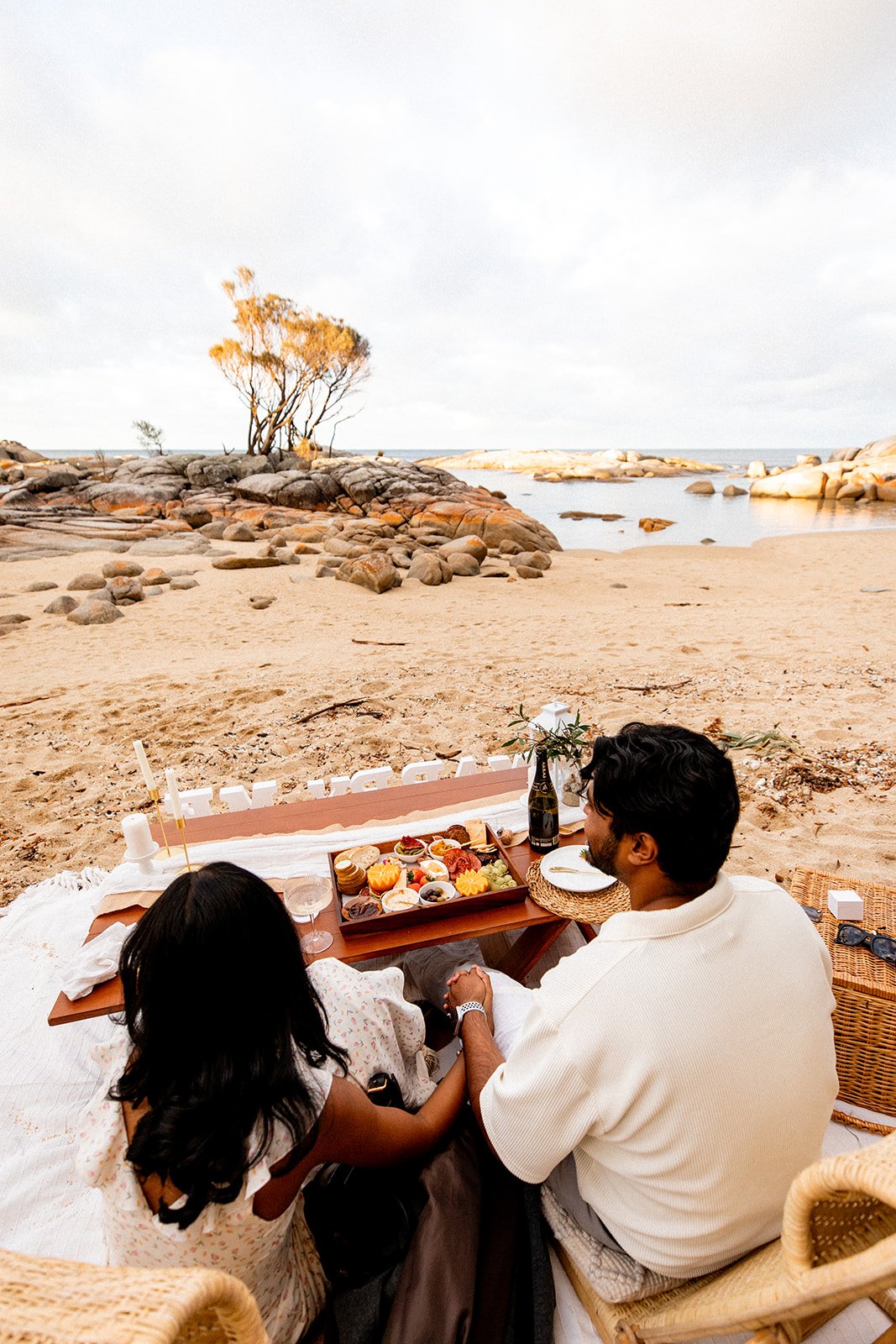 A couple and a young girl sharing a picnic on the beach with a backdrop of rocks, trees, and ocean.