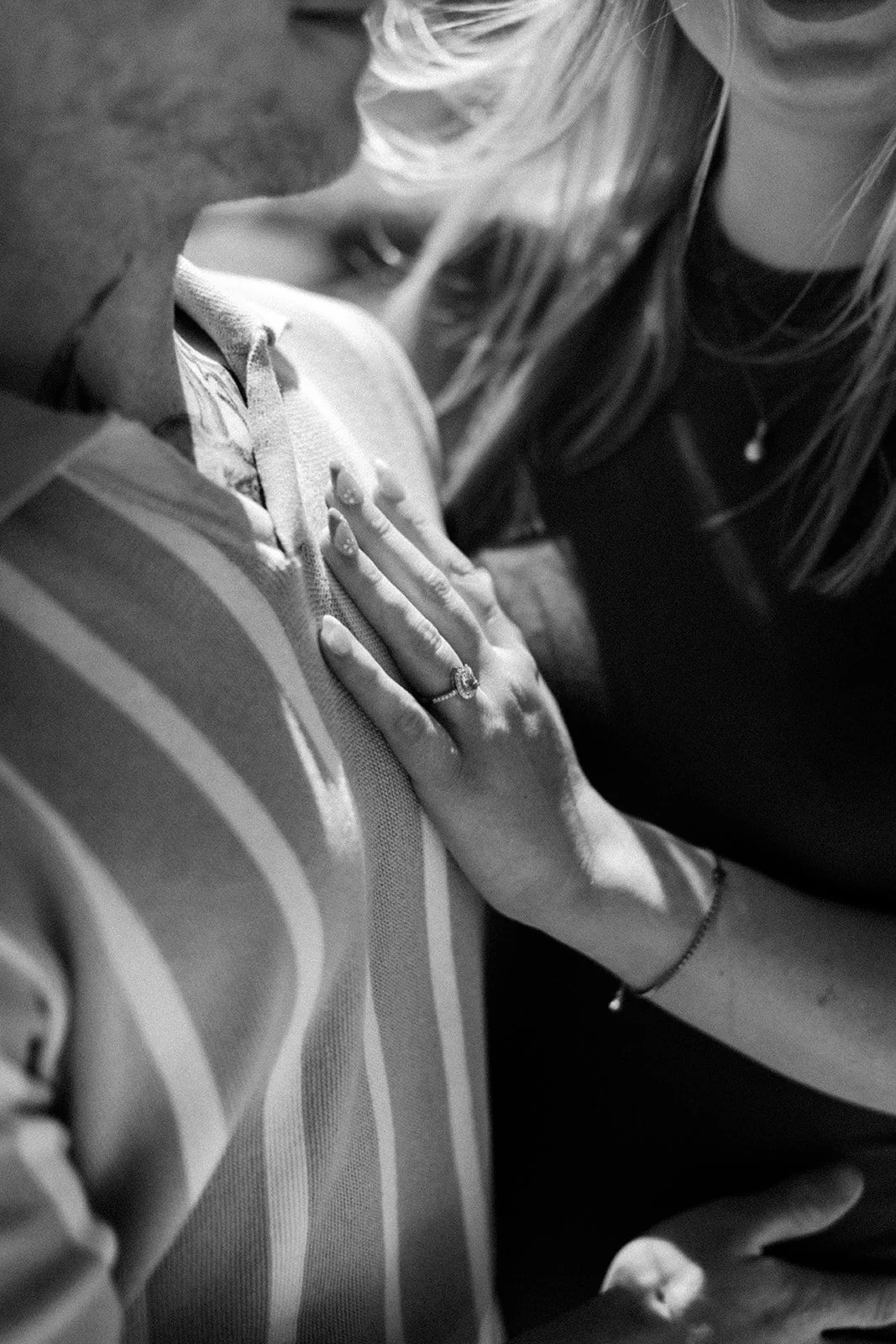 Close-up black-and-white photo of a woman kissing a man, with her hand resting on his chest, showing a ring on her finger, and another person partially visible in the background.