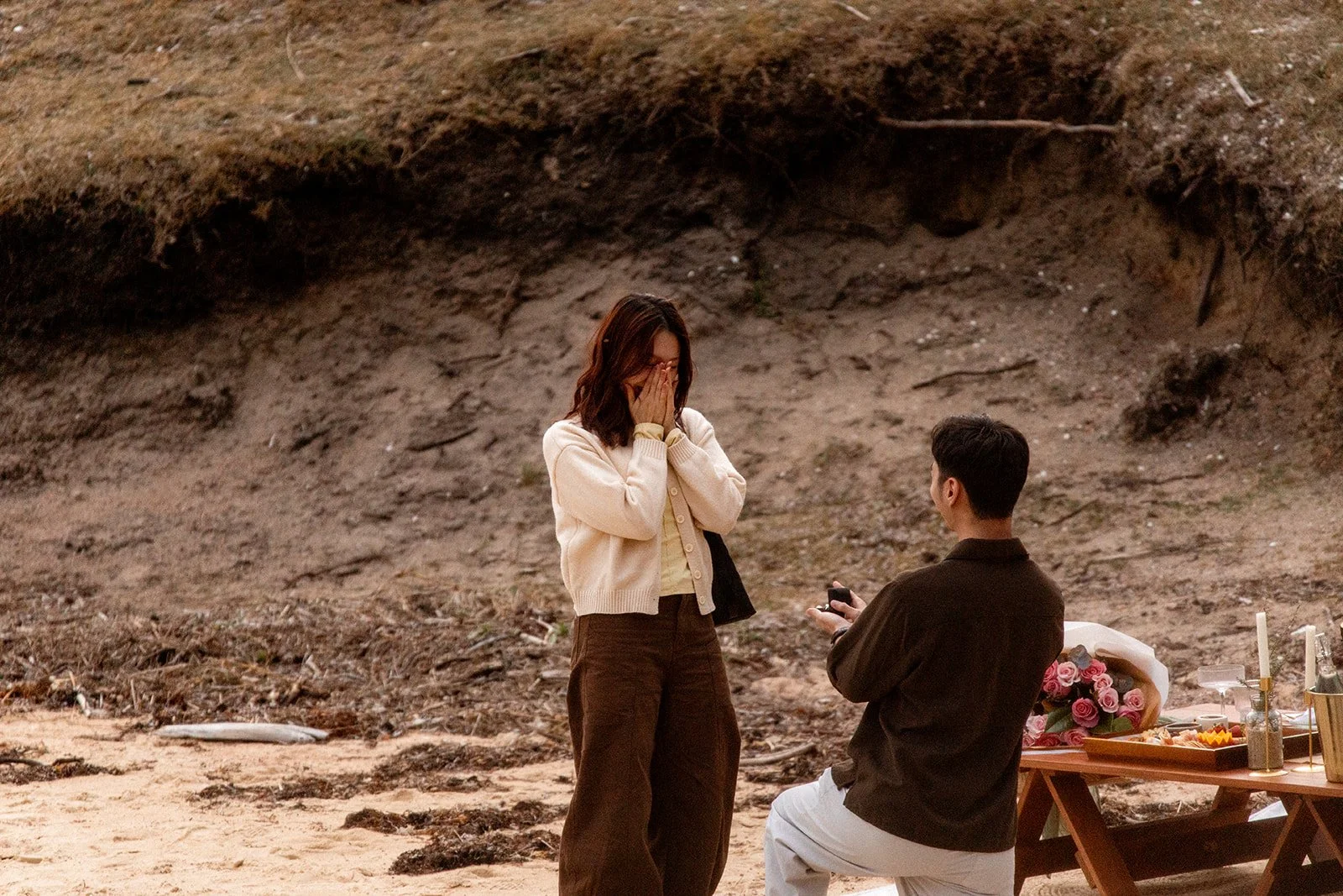 A woman appears emotional as she is proposed to by a man on a beach with a table set for a romantic occasion, including flowers and candles.