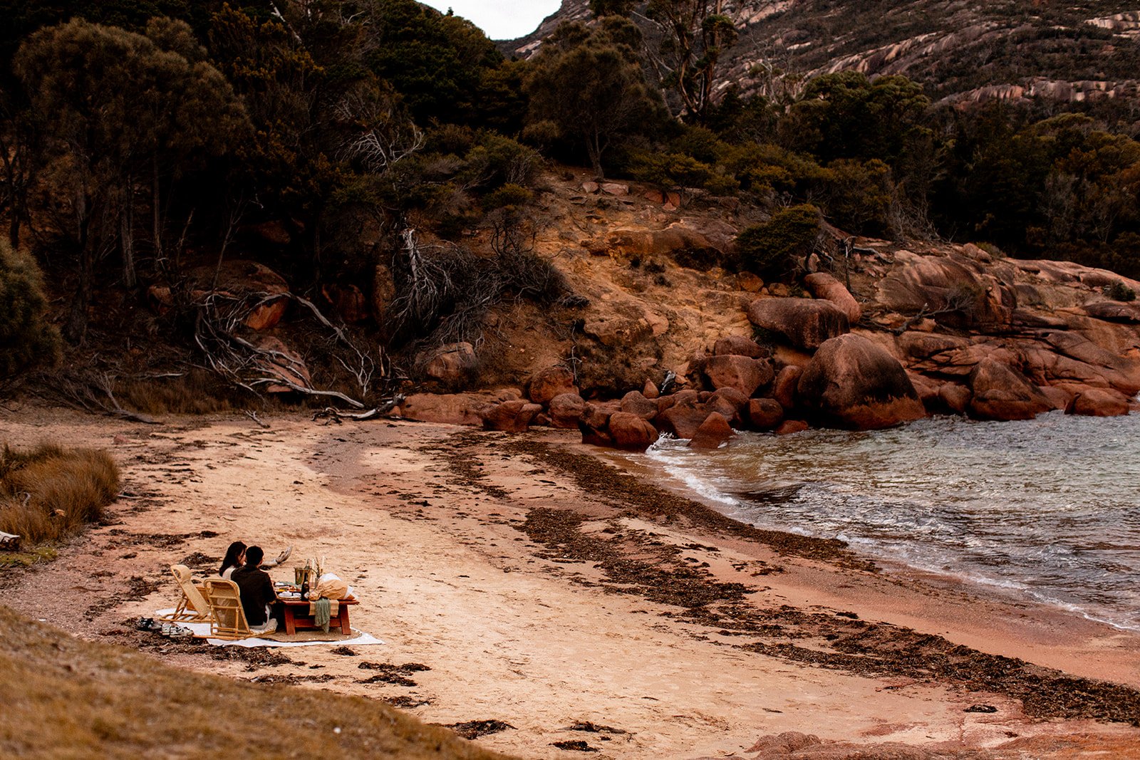 Couple sitting at a picnic table on the beach, with trees and large rocks on a hillside in the background.