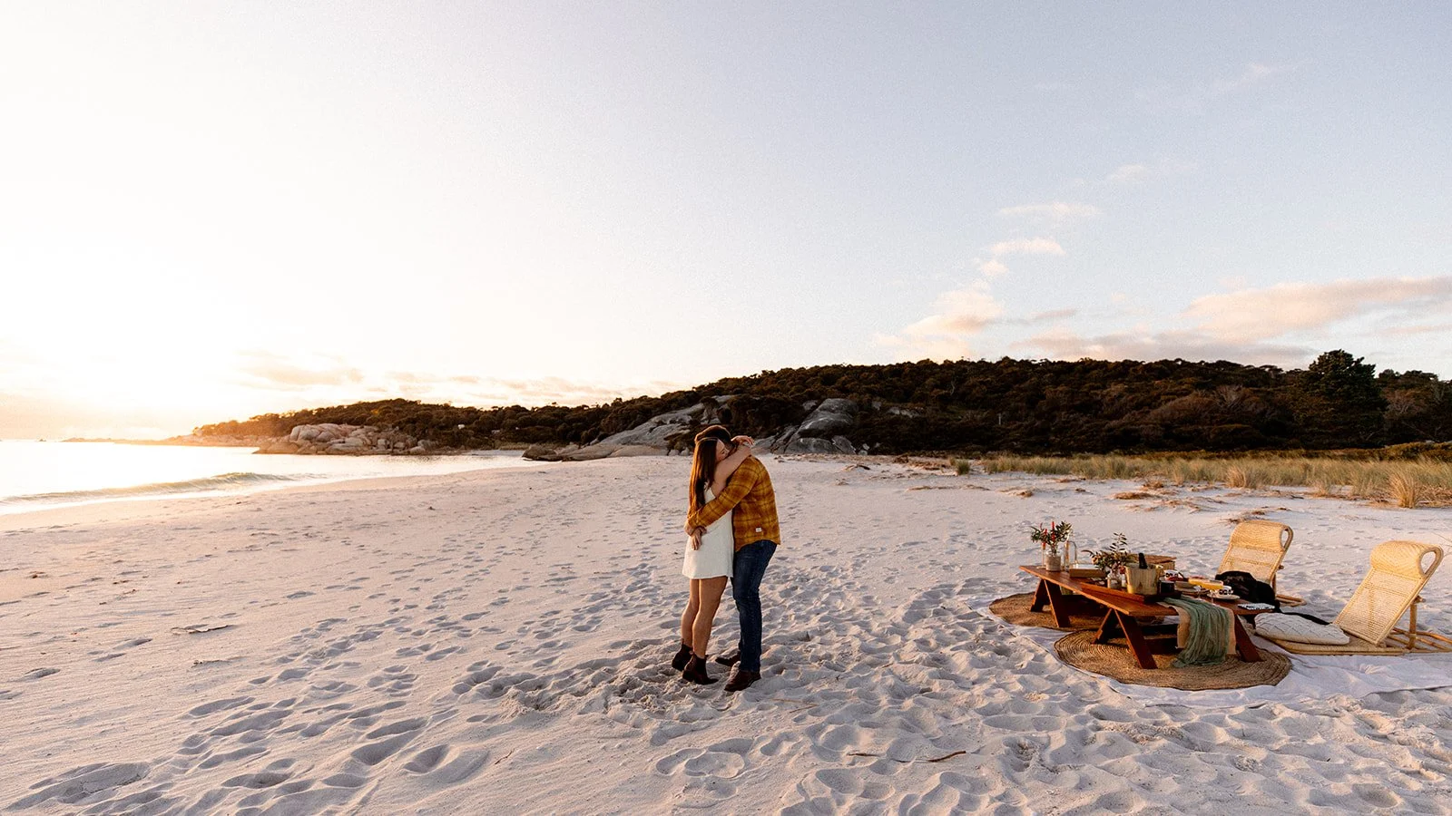 A couple hugging on a sandy beach during sunset, with a picnic setup including a low table and chairs nearby.