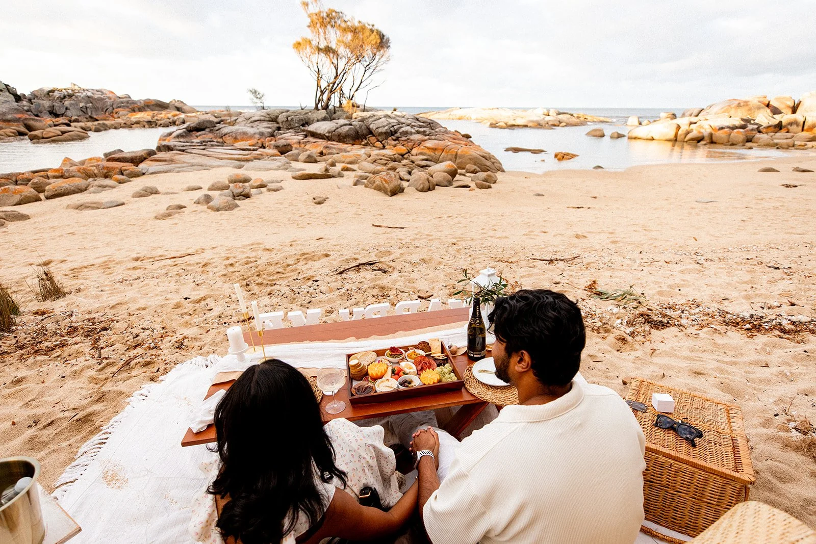 A couple enjoying a beach picnic with a view of rocks, the ocean, and a cloudy sky in a romantic location in Binalong Bay Tasmania.