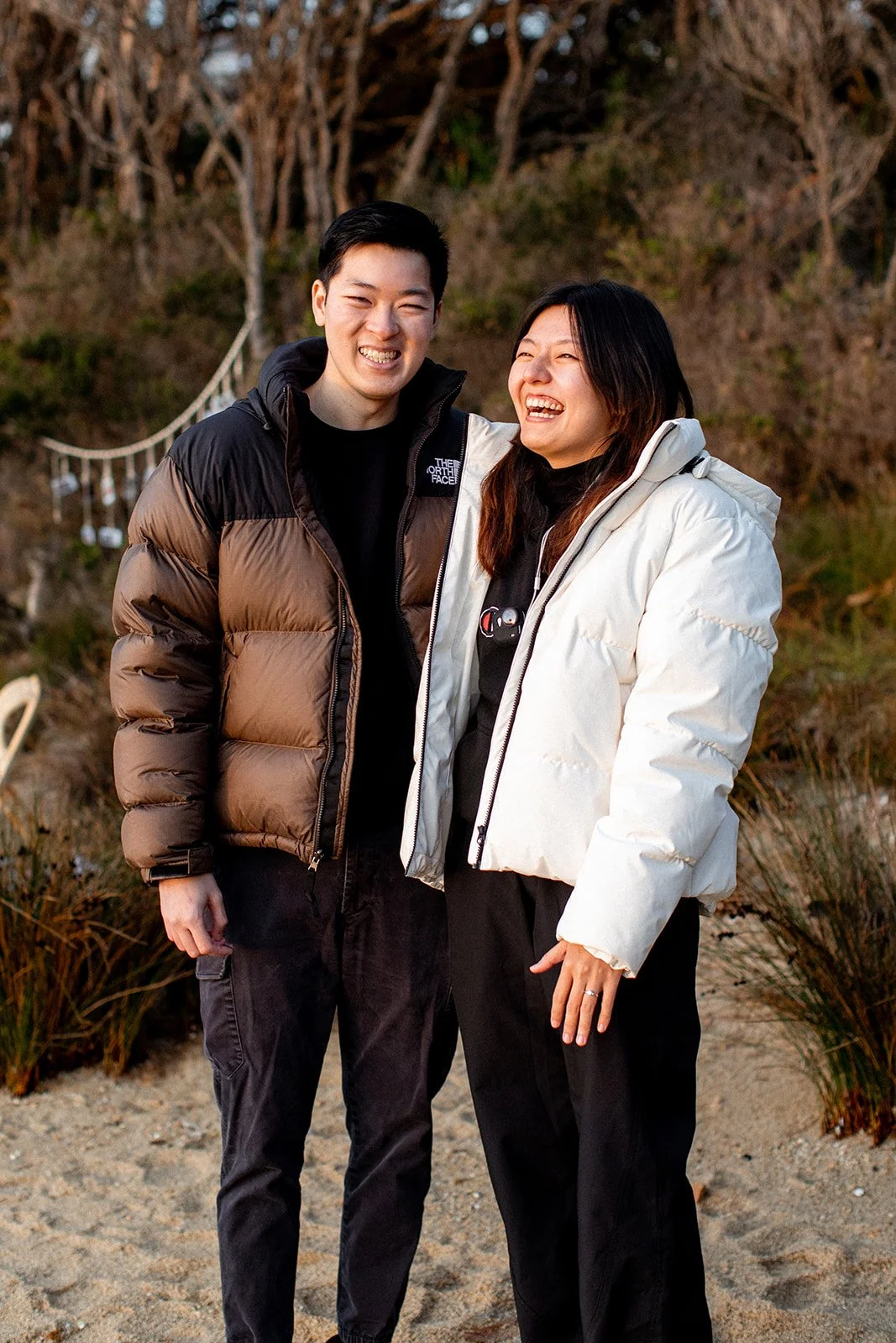 Two people, a man and a woman, standing outdoors on a beach or sandy area near rocks. They are smiling and laughing, dressed in warm jackets, with a background of trees and some decorative hanging lights.