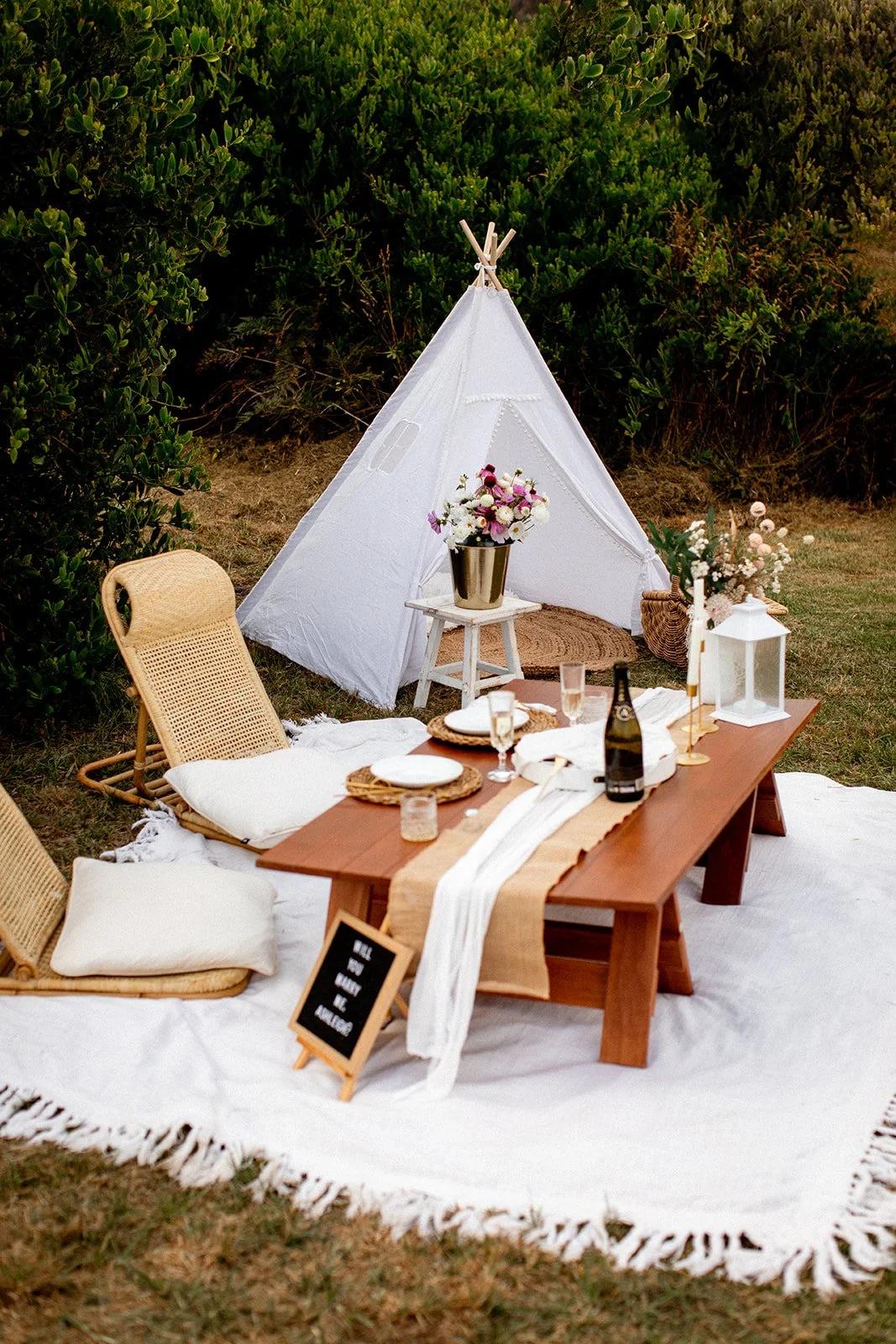 Outdoor picnic setup with a teepee tent, a wooden table with place settings, floral arrangements, and cushions on a white rug, surrounded by green foliage.