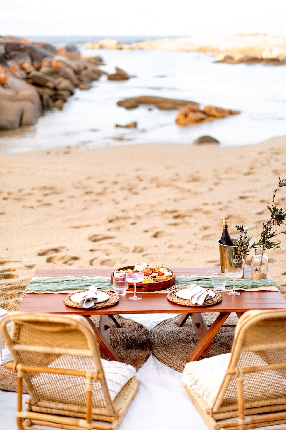 Beach picnic setup with a low wooden table, six woven chairs, plates with napkins, glasses, a centerpiece with drinks, and a platter of food, with the ocean and rocks in the background.