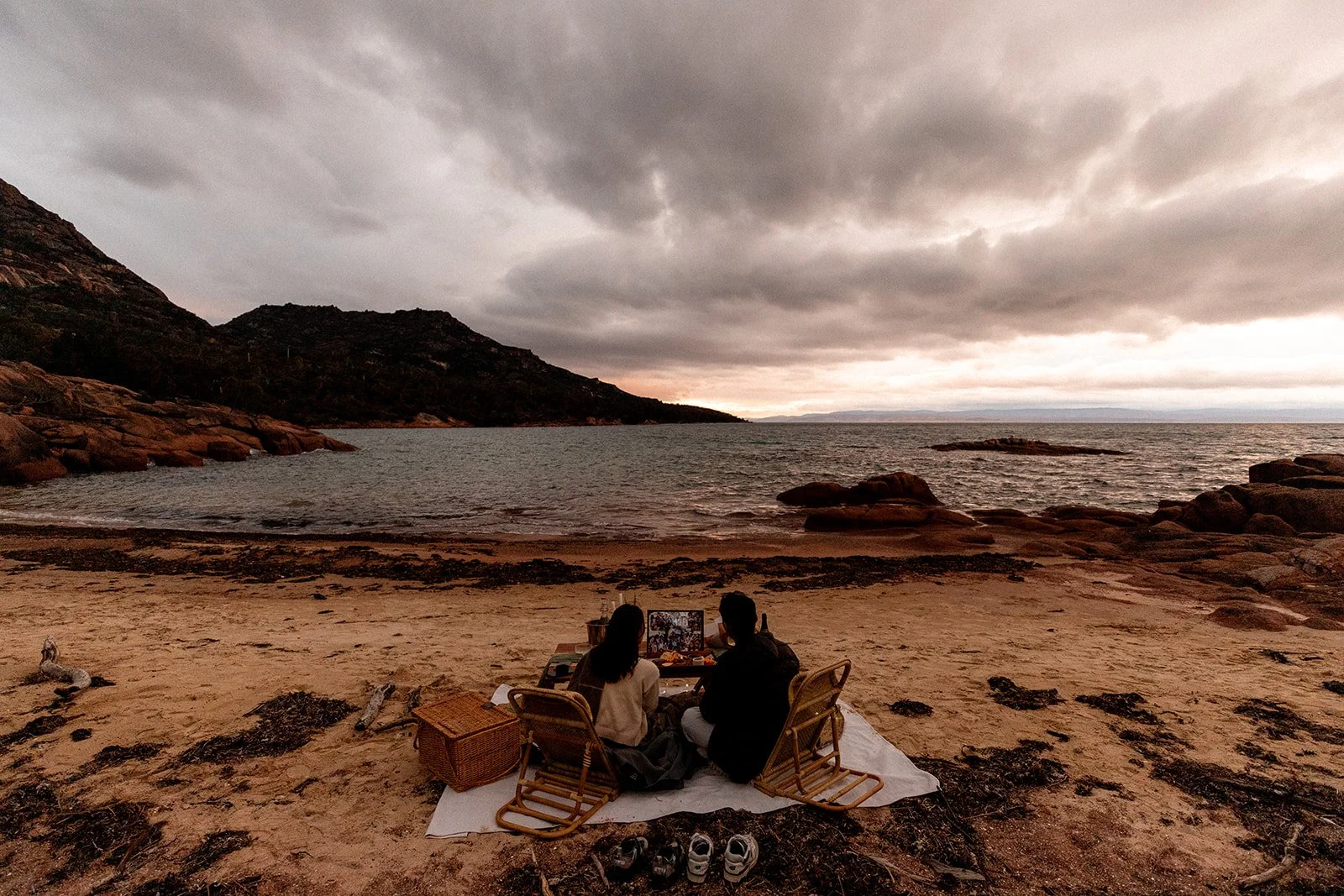 Two people having a picnic on a beach at sunset, with rocky hills and an overcast sky in the background.