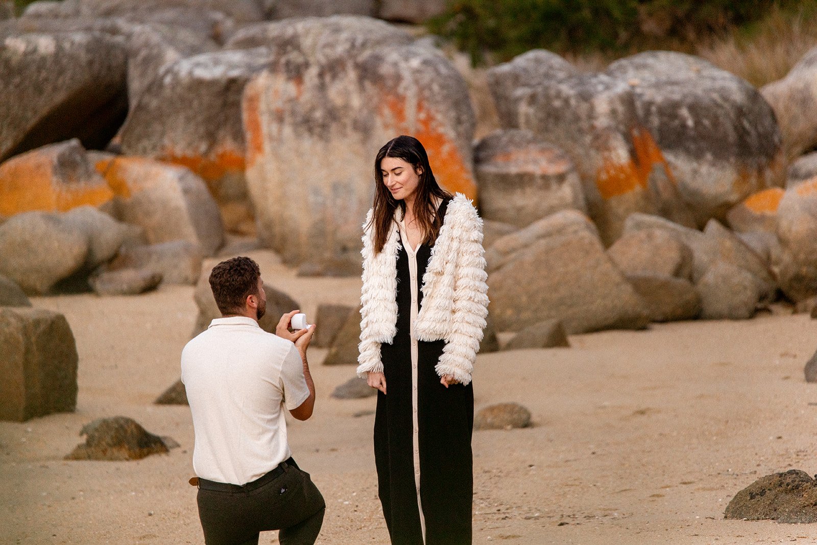 A man is proposing to o a woman in Binalong Bay with the man kneeling and holding an open ring box, and the woman reacting happily.