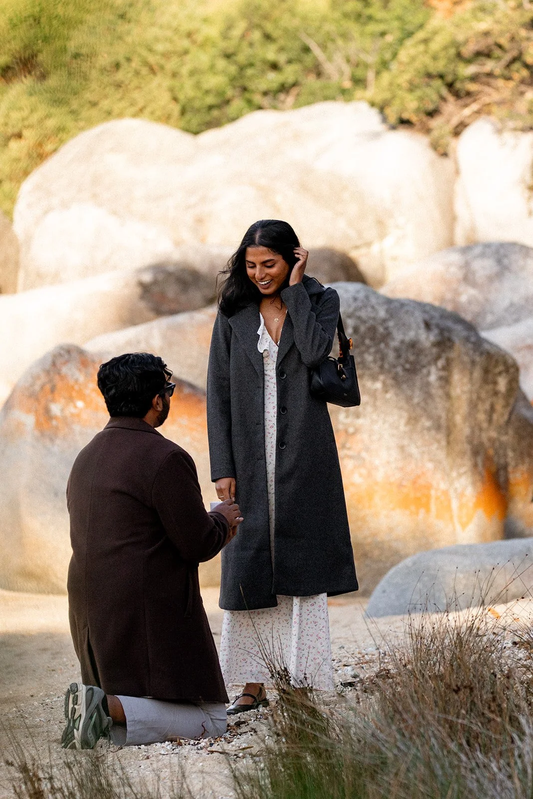A man kneeling on one knee proposes to a woman standing outdoors among large rocks. The woman is smiling and looking down at the man, who is holding her hand. The woman is wearing a long coat and a patterned dress, and the man is wearing glasses, a d