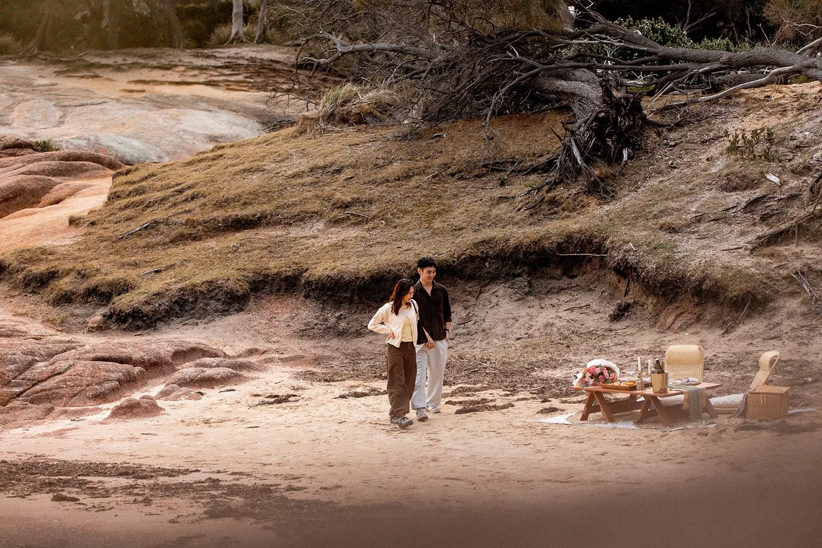 A couple walking along a sandy beach near a picnic setup with flowers, candles, and chairs, surrounded by rocky terrain and a large tree.